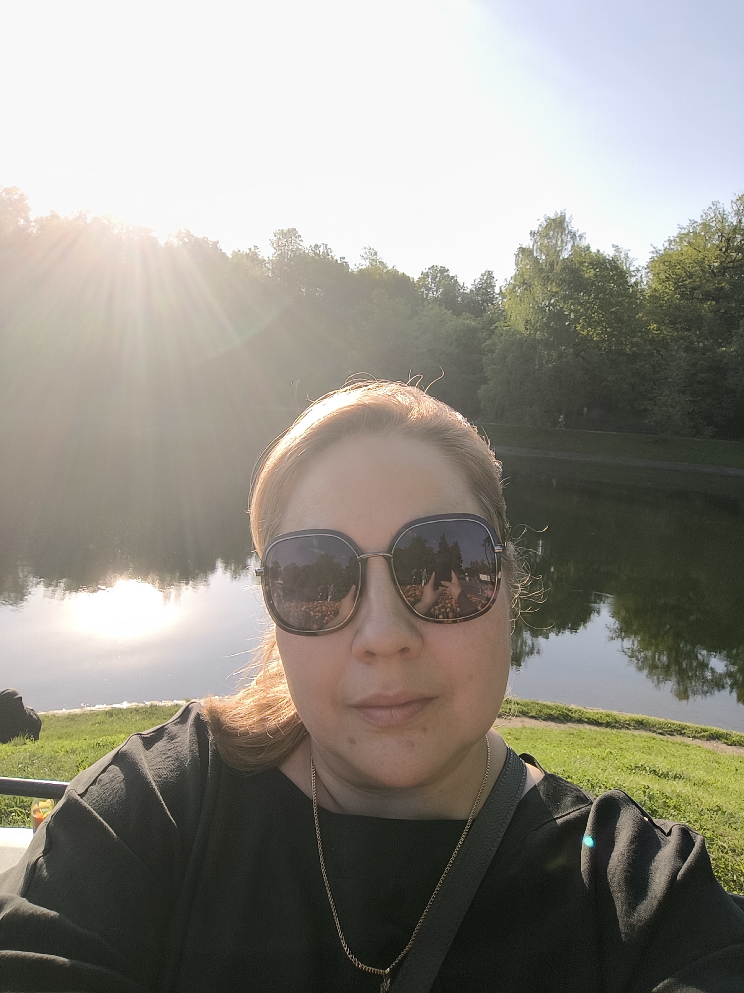 A woman wearing dark sunglasses, a black shirt, and a gold necklace is taking a selfie. The sunglasses reflect what appears to be a crowd of people and orange objects. In the background, there is a body of water reflecting sunlight. Behind the water, trees are visible under a clear sky. The photo was likely taken in a park setting in Moscow, Russia, given the person’s profile and other visual cues.