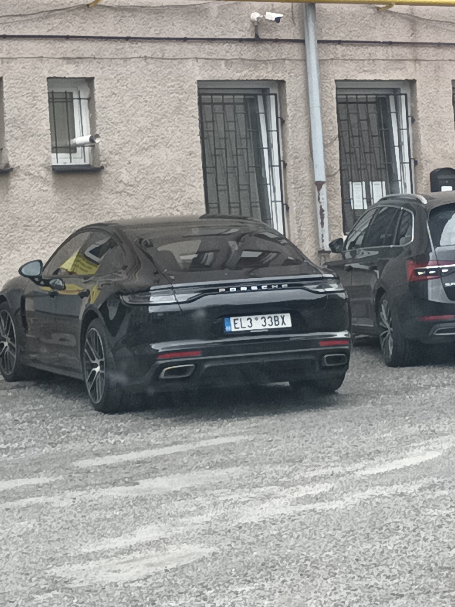 The image shows a black Porsche Panamera parked on a gravel surface in front of a building with a textured beige facade. The car has a license plate reading "EL3 33BX". To the right of the Porsche, another black car, possibly a Skoda Superb, is partially visible. The building has several windows with metal security bars and a security camera is mounted near the top of the building. A yellow pipe runs horizontally along the top of the building. The location appears to be in Svémyslice, Czechia, based on the license plate format and common car models seen in the region. The weather appears to be overcast, with a slight dusting of snow or ice on the ground, suggesting a cold climate. There are no people visible in the image.