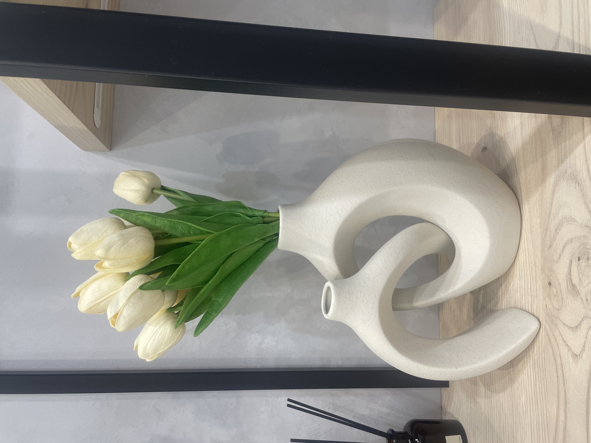 The image shows a close-up of two cream-colored, abstract ceramic vases on a light wooden shelf. The larger vase is behind and to the right of the smaller one, and both have a smooth, slightly textured finish. The larger vase is filled with a bouquet of artificial white tulips with green leaves. To the left of the vases, a dark brown glass bottle with black diffuser sticks is partially visible. In the background, a textured gray wall is present, with a vertical black metal support beam to the right. A light wood shelf is visible at the top of the frame, supported by the black metal beam. The scene appears to be an indoor display, possibly in a retail store or a home. No people are present. The location is not Saint Petersburg, Russia.