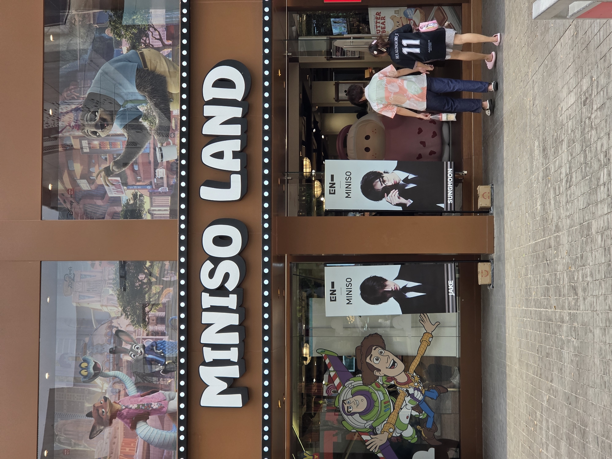 Here's a detailed description of the image:
**Scene Overview:**
The image depicts the exterior of a "Miniso Land" store. It's likely located in a busy urban area, as suggested by the paved walkway to the right.
**Storefront Details:**
* The "Miniso Land" sign is large and vertical, with white lettering outlined in black, set against a brown backdrop. The sign is bordered by a vertical row of white lights.
* The storefront windows display posters and characters from popular animated movies like *Zootopia* and *Toy Story*. There are also posters featuring EN-Miniso models.
**People:**
* Two people are visible walking near the entrance. One is wearing a floral-patterned shirt and jeans. The other is wearing a black sports jersey with the number "11" and the name "HULALONGKORN U" on the back.
**Objects:**
* The walkway is paved with grey rectangular bricks.
* A small cardboard box is visible near the entrance.
**Other Details:**
*Based on the name and imagery on the sports jersey worn by a customer, it is highly probable that the store is located in Bangkok, Thailand, near Chulalongkorn University.*