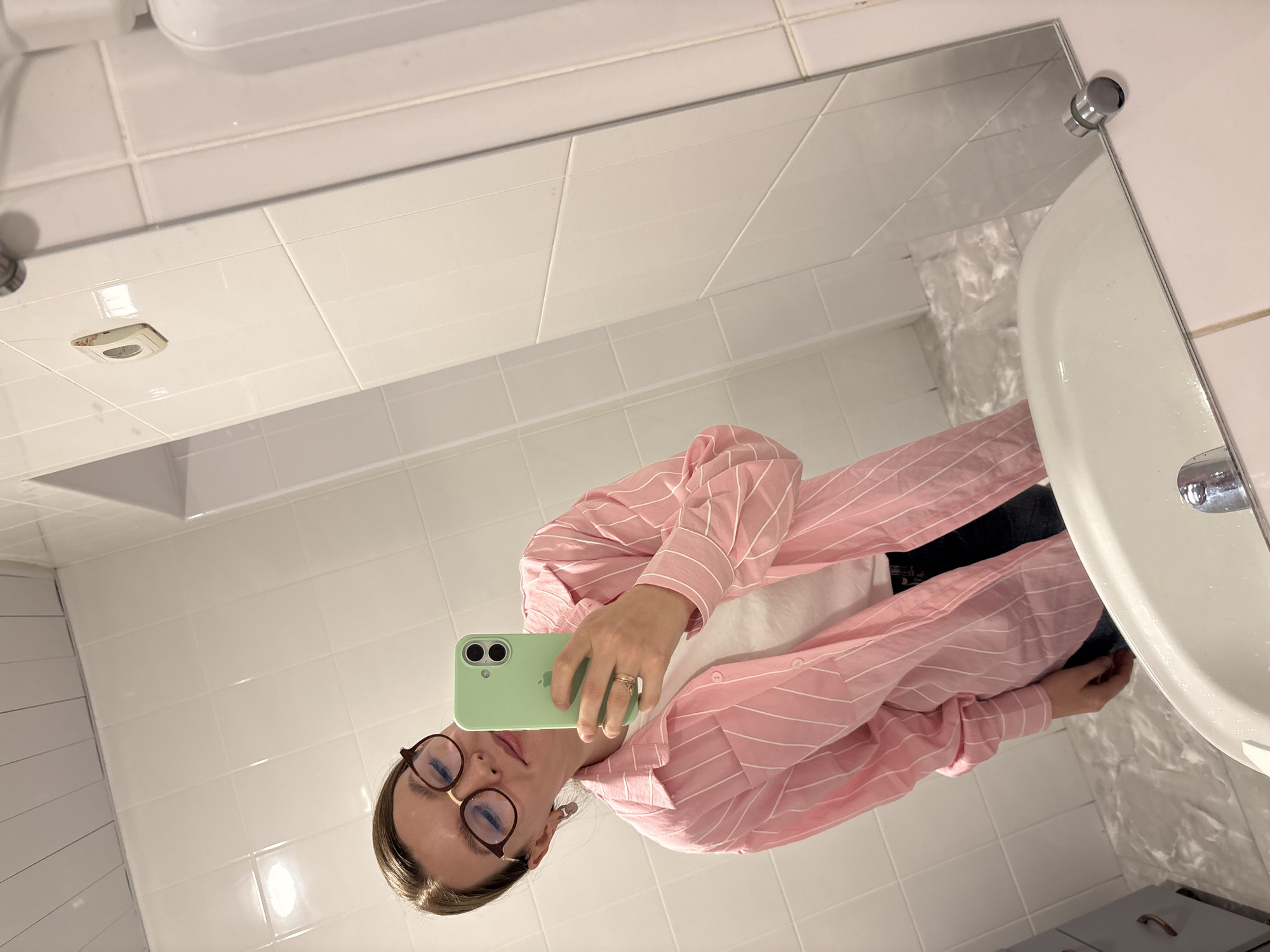 A mirror selfie depicts a person in what appears to be a bathroom setting, with the image rotated 90 degrees counter-clockwise. The subject, likely a female, is visible from the chest up, wearing a light pink and white striped button-down shirt over a white undershirt, and dark pants or jeans. She has light brown hair, wears brown-rimmed glasses, and displays a neutral expression. Her right hand holds a light green-cased smartphone (resembling an iPhone) directly towards the mirror, indicating she is using it to take the photo. A ring is visible on her right ring finger.
The background consists of white tiled walls and ceiling, typical of a functional or public restroom. A white ceramic sink with a chrome faucet is prominently featured on the right side of the frame. Below the sink, a dark-colored bin or storage unit is partially visible. An overhead recessed light fixture is also present on the tiled ceiling, contributing to the bright, artificial lighting of the space. There are no discernible outdoor elements or specific time-of-day indicators. No legible text is visible in the image. The overall setting is clean and utilitarian.