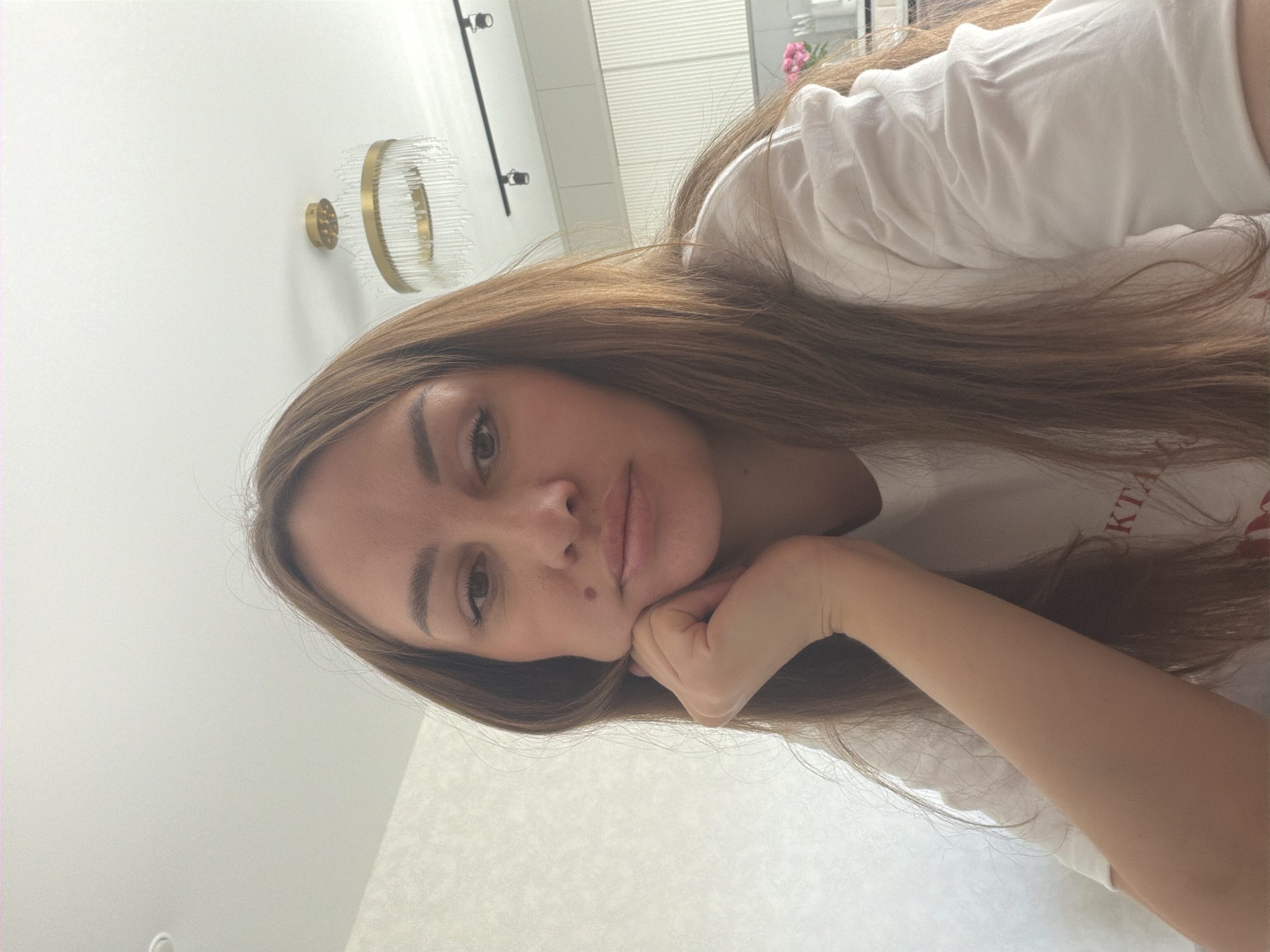 A young woman with long, brown hair is captured in a medium close-up selfie. She is wearing a white t-shirt with some red lettering visible. Her left hand is resting on her chin, and she is looking directly at the camera with a neutral expression. The background appears to be an indoor domestic setting, possibly a kitchen or dining area, with a chandelier and wall cabinets visible. The lighting suggests it is daytime. There is no indication of the city of Malinovka, Russia in the image.