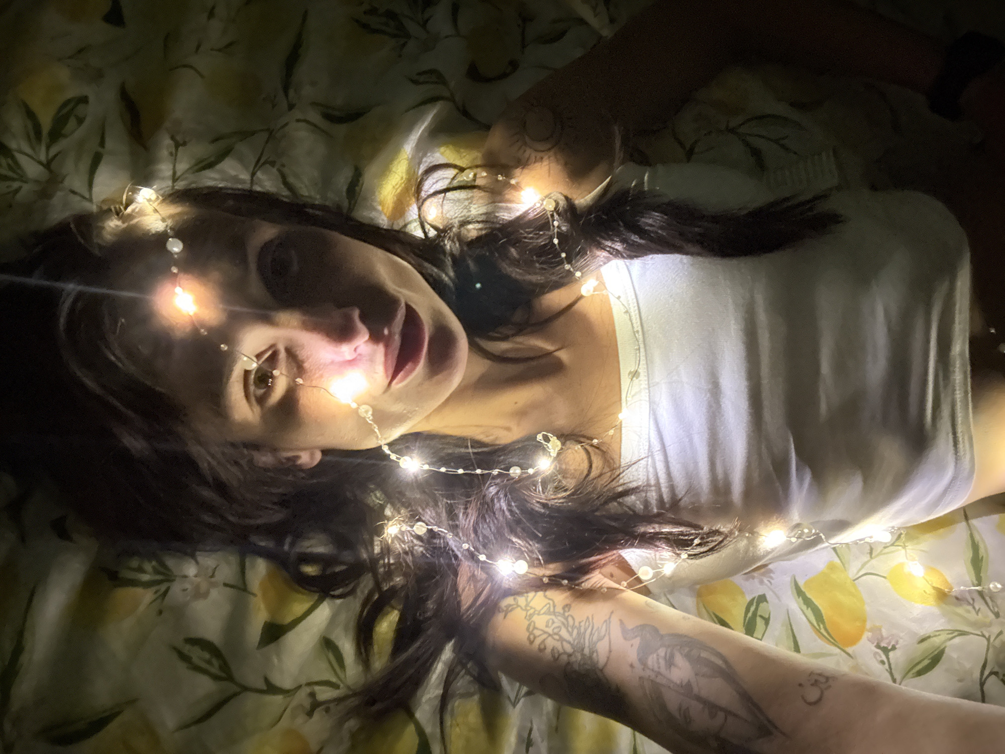 A young adult woman lies on a bed covered with a yellow and green floral print sheet. She wears a white tank top and is partially covered in strings of battery-operated lights. The woman has dark hair, fair skin, and a tattoo on her left arm. The photograph is taken at a low angle. There is no indication of this photo being in Pleven, Bulgaria.