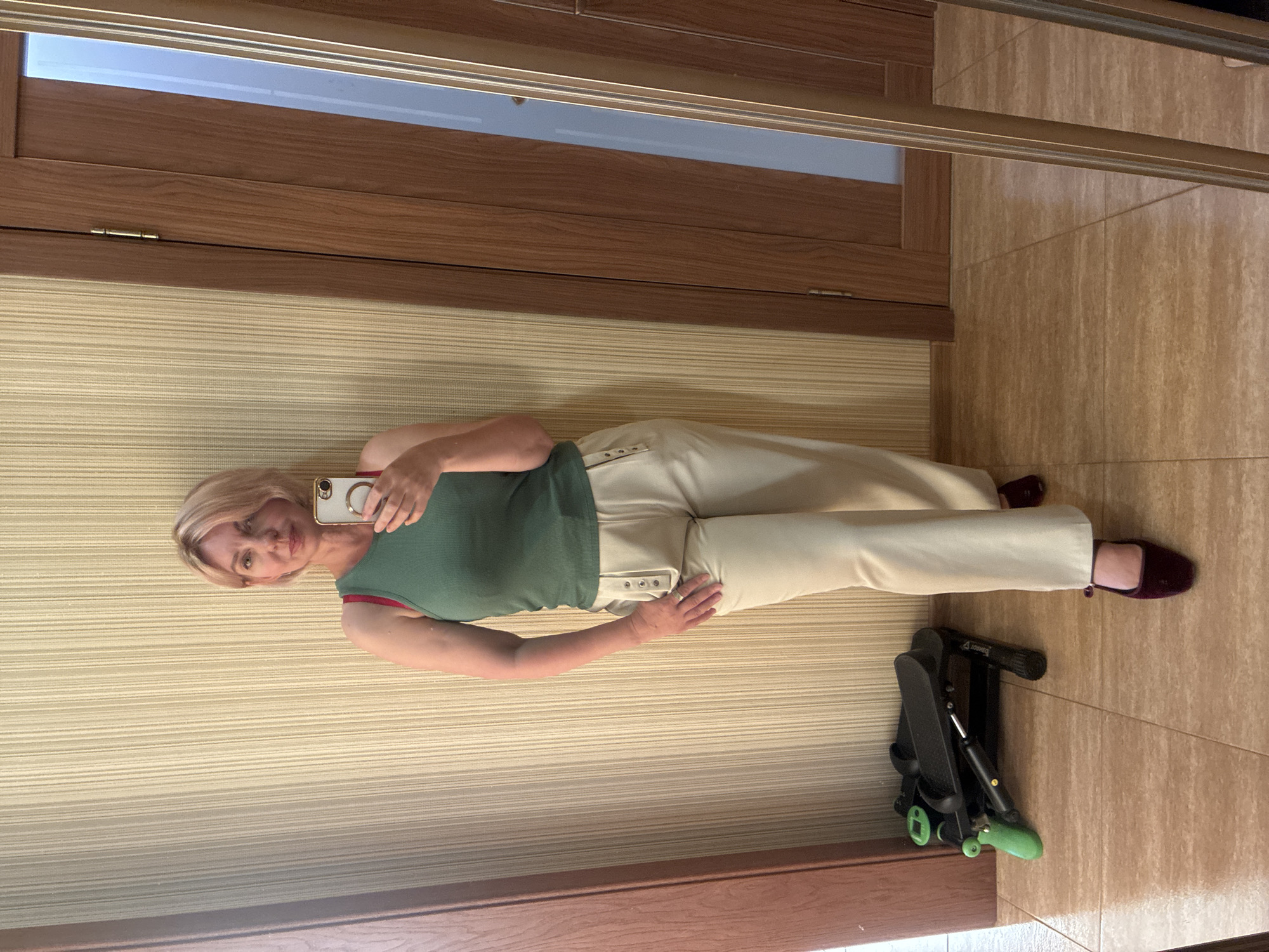 An image taken in Sochi, Russia, depicts a fair-skinned woman in her 40s or 50s, standing in an interior room and capturing a mirror selfie. She has short, light blonde hair and is looking directly at the camera with a neutral expression. The woman is dressed in a green ribbed tank top, with a red bra strap visible on her left shoulder. She wears light beige wide-leg trousers featuring decorative buttons and a loop design on the front hip pockets. Her footwear consists of dark reddish-purple velvet flat shoes with an instep strap, worn over light-colored socks. She holds a light-colored smartphone with a prominent gold ring stand in her right hand, angled to photograph her reflection.
The background features a light beige wall covered in vertically striped wallpaper. To the left, on a light brown tiled floor, a compact black and green exercise stepper is positioned. To the right, a medium-brown wooden door with visible grain and a brass hinge is present, reflecting a frosted glass panel. A tall, gold-framed mirror on the right edge of the frame reflects parts of the room, including the door. The floor is composed of large, square, light brown tiles with dark grout lines.