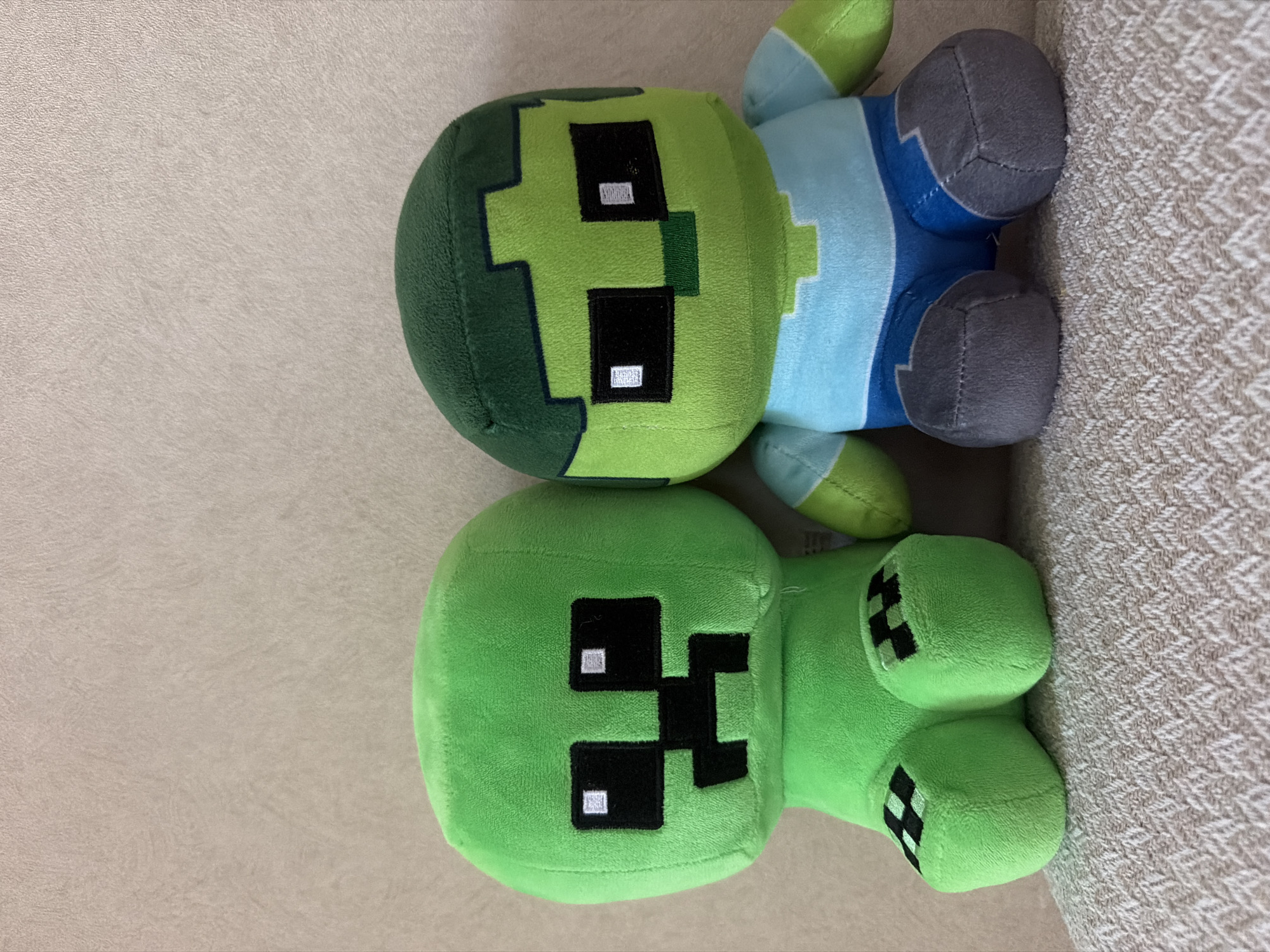 Two Minecraft plush toys, a green Creeper and a green and blue Zombie, are positioned side-by-side on a textured, light-colored surface, likely a couch or chair, against a neutral, textured wall. The Creeper, on the left, is entirely green with a pixelated black face featuring two square eyes and a square mouth. Its feet are green with black checkered patterns. The Zombie, on the right, has a green, pixelated head with black square eyes and a green nose. It wears a light blue shirt and blue shorts. Its feet are gray. The lighting is even and appears to be natural or from an overhead source, casting no significant shadows. The background wall has a subtle, regular texture. The scene is indoors and static, with no discernible action or interaction occurring. The location is not specified as Bishkek City, Kyrgyzstan.
