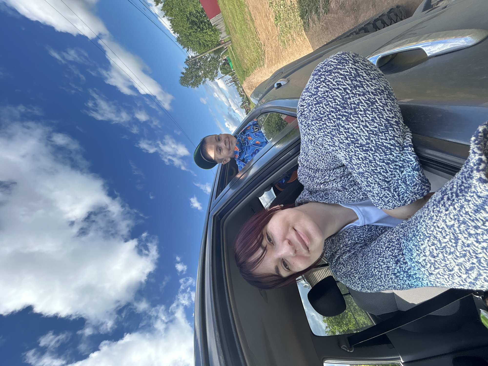 A woman and a child are in a car on a sunny day. The woman is in the foreground, wearing a blue and white patterned sweater and white shirt. She has dark hair with purple undertones and is looking at the camera with a slight smile. Her legs are visible, also wearing the patterned sweater. The child is in the backseat, leaning out of the window. He is wearing a blue t-shirt with an orange pattern and a green baseball cap. He is smiling and looking towards the camera. The car's interior is visible, including a seatbelt and a side mirror. Outside the car, the sky is bright blue with fluffy white clouds. There are trees and a field visible in the background, suggesting a rural or semi-rural setting. Power lines are also visible against the sky. The overall impression is a candid, happy moment captured during a car ride. The time of day appears to be daytime, likely late morning or afternoon, given the bright sunlight and clear sky. The weather is sunny and pleasant. The location context provided is Foky, Russia. There is no visible text in the image.