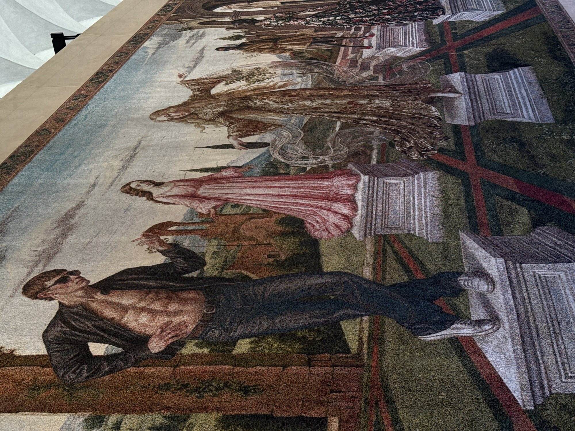 This is a tapestry depicting an urban scene. A shirtless man wearing sunglasses, a black jacket, and dark pants lies horizontally across the foreground. Above him stands a woman in a red dress, and further up is an ethereal, winged figure. At the top of the tapestry are several figures, including a man in armor. The scene is rendered in muted colors, with green, brown, and red tones dominating. Architectural elements, such as pillars and archways, suggest an urban setting. There is no verifiable information linking this image to Milan, Italy.