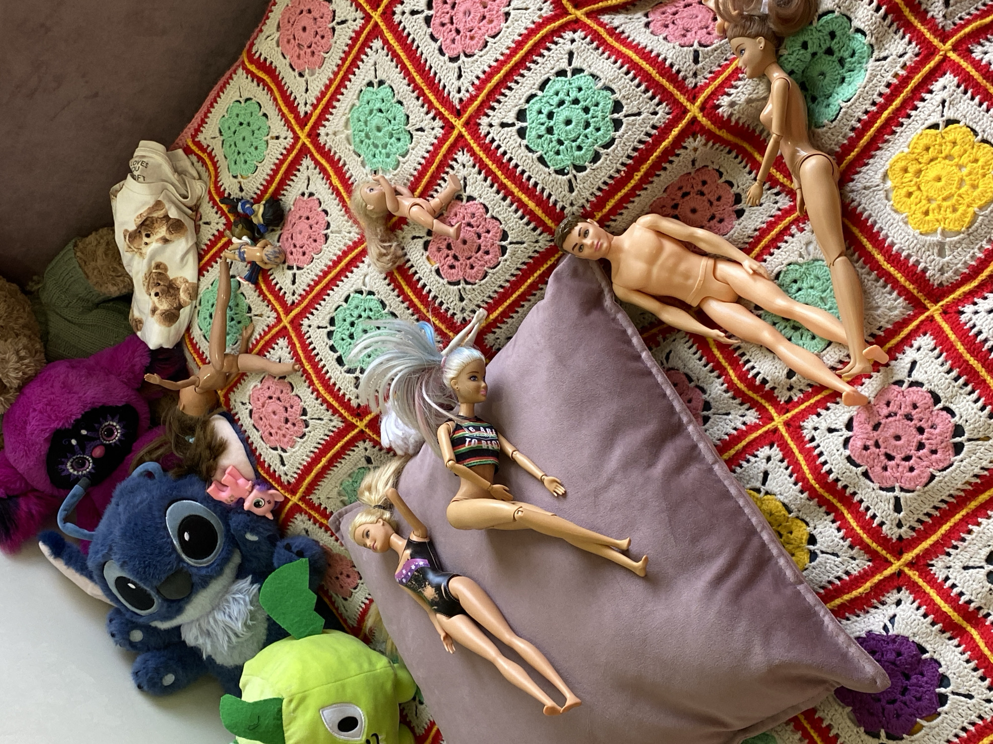 An overhead shot captures a colorful arrangement of dolls and plush toys resting on a vibrant granny square crochet blanket and a light purple pillow. The blanket features a distinctive pattern of granny squares with centers in pink, light green, yellow, and purple, outlined in white, and interconnected by a bold red and yellow grid.
The subjects include several Barbie-type dolls and one Ken-type doll, most of which are nude, while some wear partial clothing. One Barbie is dressed in a striped crop top, and another in a dark swimsuit, with one doll notably having long, light-colored hair with highlights. Smaller plastic dolls are also scattered on the blanket.
Among the plush toys, a large blue Stitch character from Disney's Lilo & Stitch is prominent in the lower left, alongside a fuzzy purple creature with large eyes and a bright green, blocky plush toy. Other soft items present are a crumpled white garment with a teddy bear print, on which the partial text "LOVES OU FT" is visible, a green knitted item, and some brown fur-like material. The scene depicts a casual collection of toys in what appears to be an indoor setting, likely a bed or couch.
Location: Votkinsk, Russia.