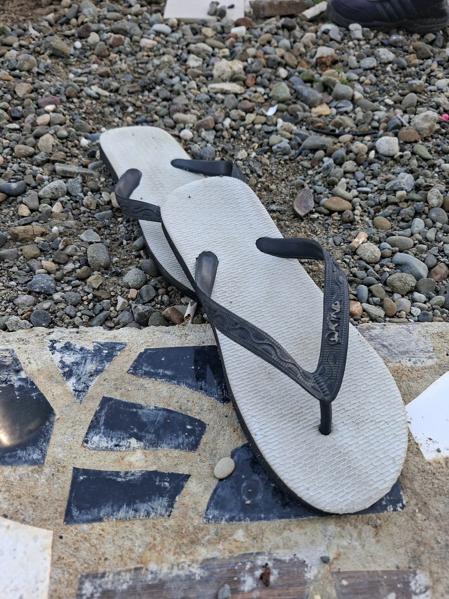 A pair of light grey flip-flops with black straps lies on a textured surface composed of small, multi-colored pebbles and sand. The flip-flops are positioned diagonally, with the soles facing upwards. The straps have a decorative pattern and a raised brand name which appears to be "Havaianas." The textured surface extends to the bottom left and center of the image, where there are also sections of a mosaic-like pattern with dark rectangular and triangular tiles. In the background, more pebbles and some darker debris are visible. The scene appears to be outdoors, possibly in a coastal or riverside area. The image quality is decent, with good detail and color balance. The lighting suggests it is daytime, though possibly overcast. No people are visible in the image.