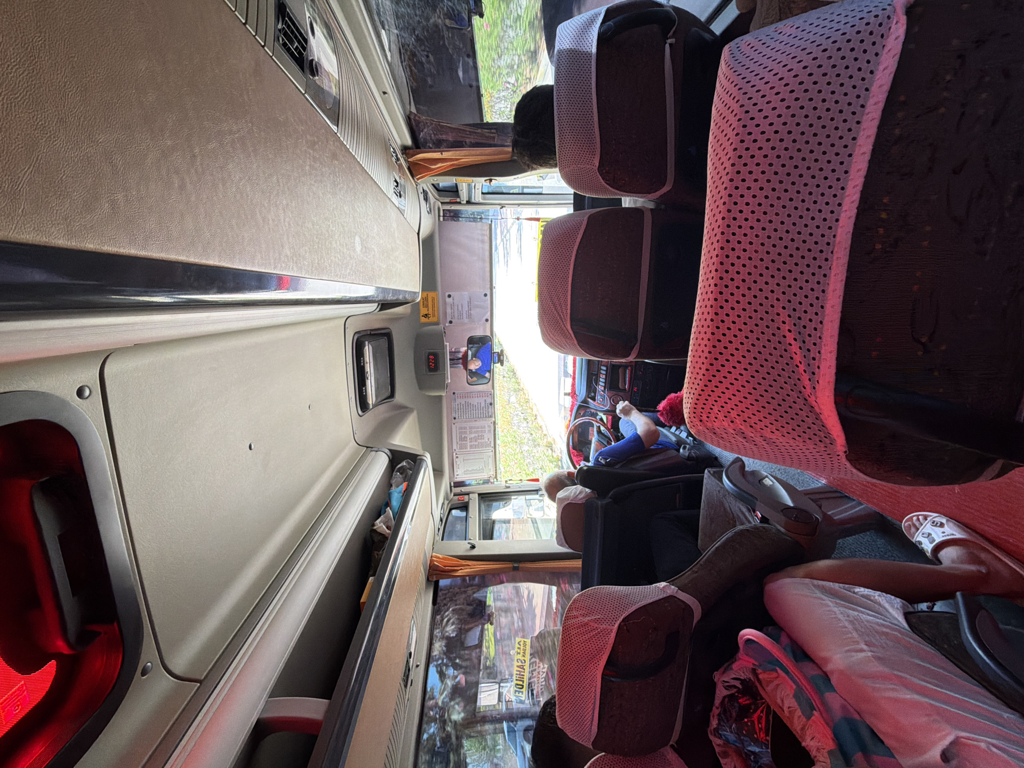 The image captures the interior of a bus, likely in Kemer, Turkey, given the presence of an outdoor scene visible through the windows that suggests a warm climate with lush greenery. The perspective is from a passenger seat, looking forward towards the driver's area and the front of the bus.
In the foreground, the backs of several seats are visible, covered with white fabric with a pinkish hue from the ambient lighting. One seat in the lower left corner has a person's legs and feet resting on it, with a patterned sandal visible on the right foot. A bag with a colorful pattern is also present on this seat.
Further forward, the driver, an older man with gray hair, is seen operating the bus. He is wearing a blue shirt and is holding the steering wheel. A digital clock on the rearview mirror displays "12:28". Above the driver, a sign with Turkish text is visible, along with an orange curtain.
The bus's interior features overhead compartments and lights, with a reddish light illuminating the front of the ceiling. Through the windows on the sides and front, an outdoor environment with trees, foliage, and a road is visible. A white car is seen on the road outside, and a yellow sign with reversed text can be observed, potentially indicating a business or a road direction. The overall impression is of a bus journey during daylight hours.