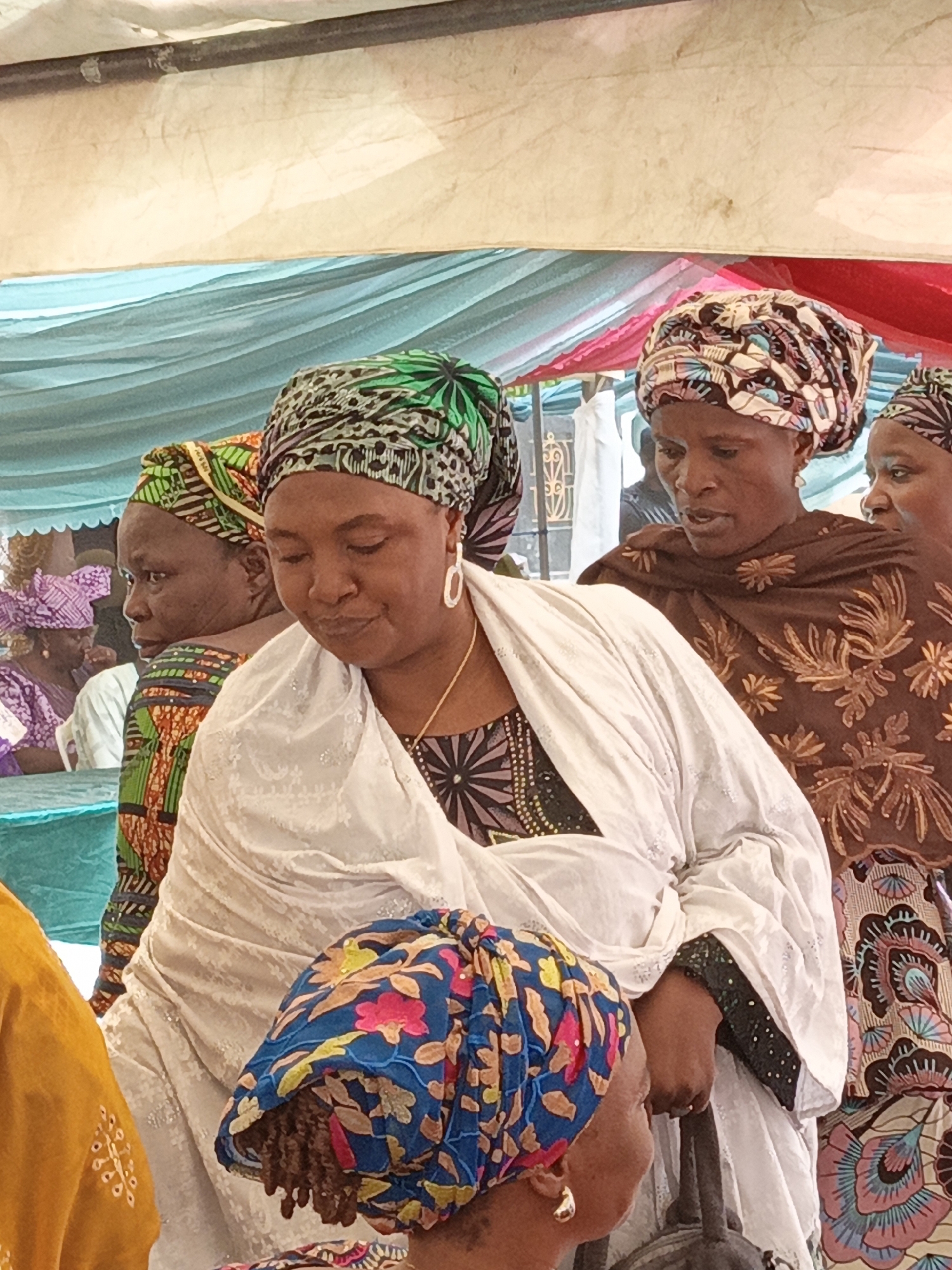 This image captures a vibrant social gathering of women under a decorated tent or canopy in Sabon Gari West, Nigeria. The scene is set outdoors or in a semi-open venue, indicated by the colorful fabric drapes overhead in shades of light blue, red, and beige, suggesting a festive ambiance during daylight hours.
The main subject, prominent in the foreground, is a woman wearing a white, intricately embroidered shawl draped over a dark, patterned top. Her head is elegantly wrapped in a colorful green, black, and white fabric, and she accessorizes with large white hoop earrings and a gold chain necklace. Her gaze is directed downwards, suggesting a moment of contemplation or observation. She appears to be holding a dark, indistinct object with her right hand.
To her left, another woman is partially visible, dressed in a vibrant green, orange, and black patterned fabric, including her headwrap, and looking towards the left of the frame. To the right of the central figure, another woman is seen wearing a brown shawl adorned with intricate patterns and a matching headwrap. Her mouth is slightly open, hinting at a moment of speech or engaged listening.
In the lower foreground, a fourth woman, with braided hair, is partially shown, wearing a striking blue headwrap featuring pink and yellow floral designs. Several other women, some wearing purple headwraps, are blurred in the background, emphasizing the communal nature of the event. The clothing styles, characterized by rich patterns and headwraps, are traditional to the region. There is no visible text in the image. The atmosphere suggests a cultural celebration, ceremony, or community meeting.