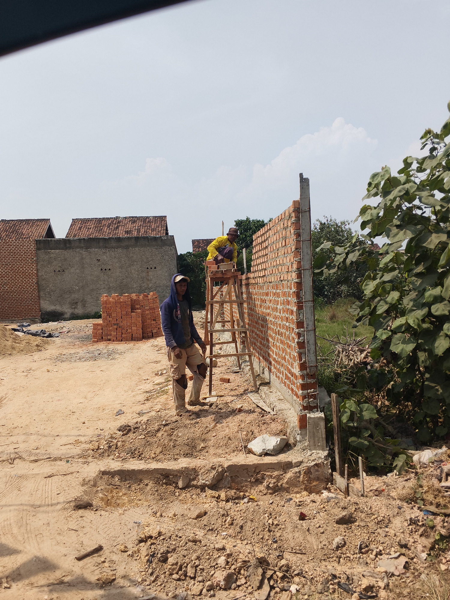 Two construction workers are observed working on a brick wall in a semi-urban environment, possibly on the outskirts of Bandar Lampung, Indonesia. The worker in the foreground, wearing a blue hooded sweatshirt, tan pants with knee pads, and a baseball cap, stands on the ground, looking towards the camera. Behind him, another worker, clad in a yellow shirt and dark pants, is positioned on a wooden ladder, actively engaged in laying bricks on the wall. The wall itself is constructed from red bricks and appears to be partially completed. In the background, several unfinished brick buildings with tiled roofs are visible, along with stacks of bricks and construction debris. The ground is a mix of dirt and gravel, and the scene is set against a backdrop of trees and a hazy sky. The overall impression is one of ongoing construction activity in a developing area.