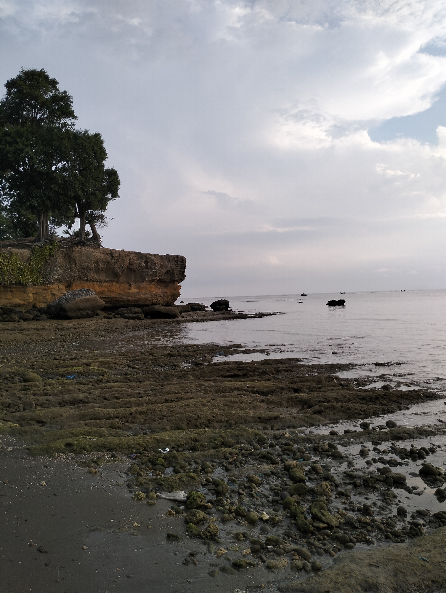 The image shows a coastal scene in Lhokmanee, Indonesia. A cliff topped with trees dominates the left side of the frame. The cliff appears to be made of layered rock and soil. The foreground consists of a rocky beach, partly submerged by the sea. In the distance, the sea is visible with a few small boats scattered across the horizon. The sky is overcast with grey and white clouds. There are no people visible in the image.