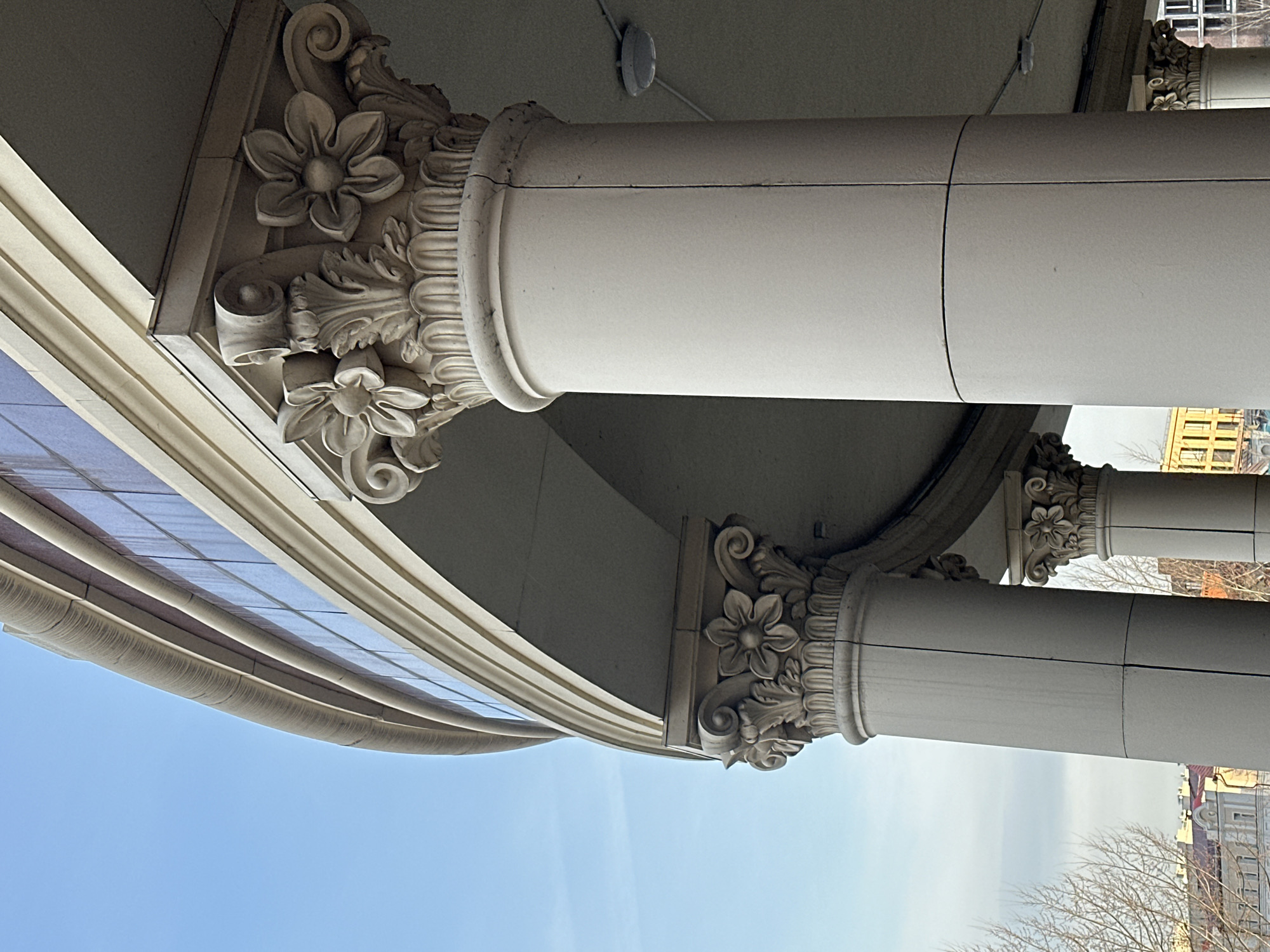 The image shows a low-angle view of architectural details, specifically of several white columns with ornate floral capitals and bases. The columns support a curved structure with multiple layers or ledges that appear to be part of a building's facade. The sky is visible in the lower portion of the frame. In the background, the tops of other buildings and bare tree branches suggest an urban environment. Based on the architectural style, it appears to be a historical or culturally significant structure, possibly in the city of Kazan, Russia.