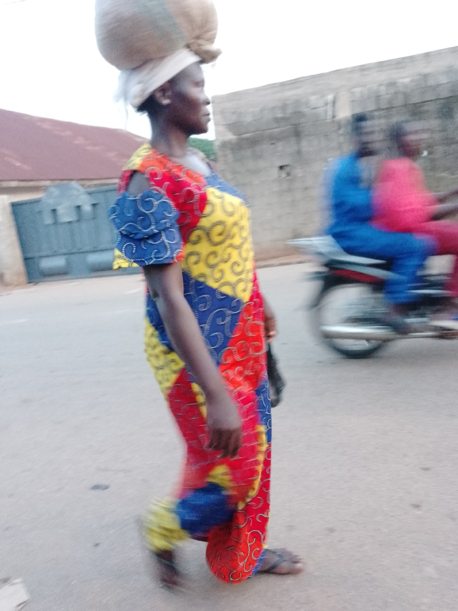 A woman with a sack on her head walks along a street in Akwanga, Nigeria. She wears a brightly colored patchwork dress. In the background, two people ride a motorcycle. Buildings line the street. The scene appears to be photographed during the day.