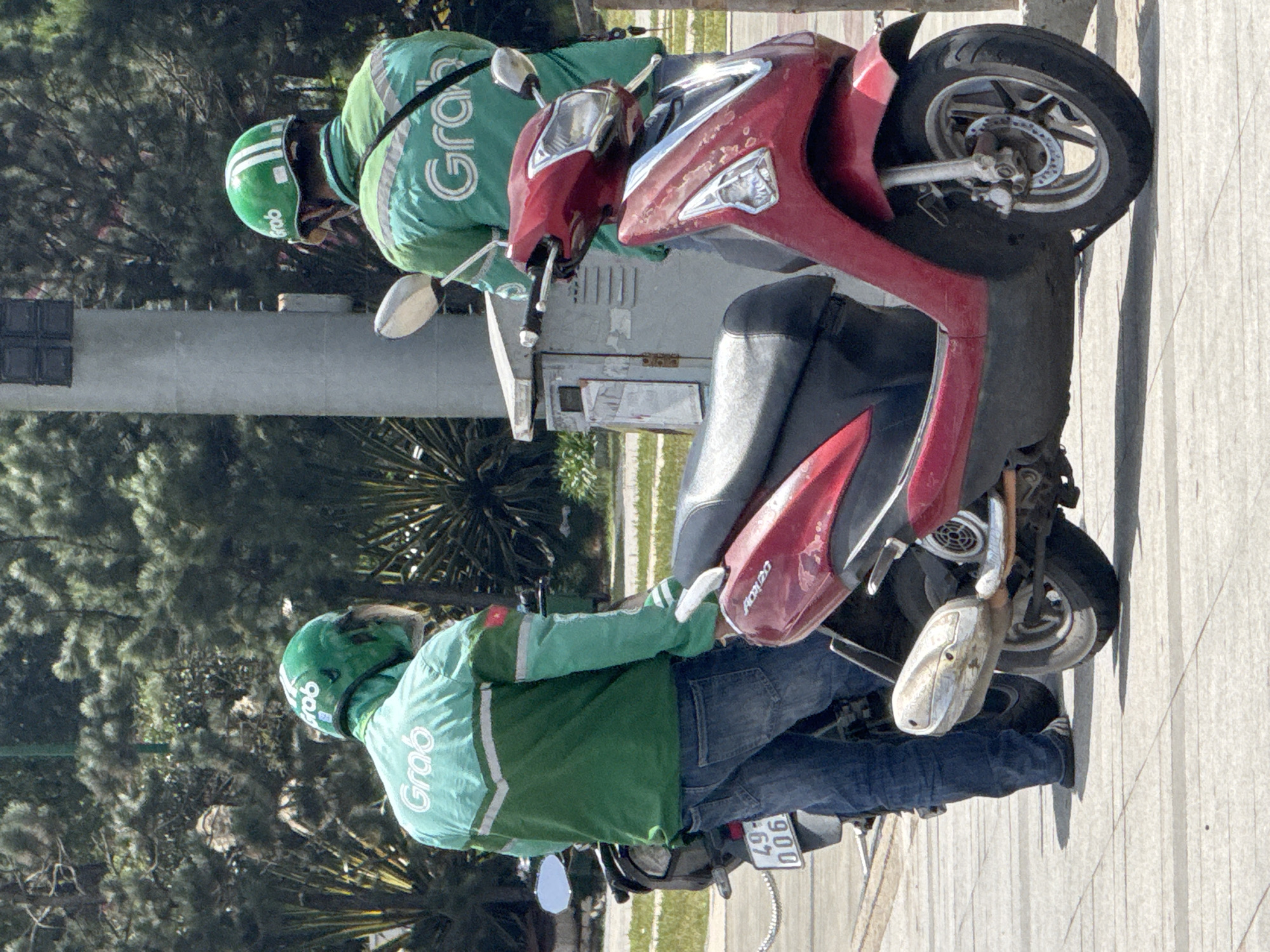 Two Grab delivery drivers, wearing green helmets and jackets with the Grab logo, are standing next to their scooters on a sidewalk in Da Lat, Vietnam. The driver on the left is facing away from the camera, wearing blue jeans and a green jacket. The driver on the right is also facing away from the camera, wearing a black crossbody bag over their green jacket. A red scooter is parked in the foreground, and a black scooter is partially visible behind the driver on the left. The background consists of trees and a light pole. The sidewalk is paved with square tiles. The scene appears to be a typical street view in Da Lat, a city known for its scenic landscapes and vibrant atmosphere.