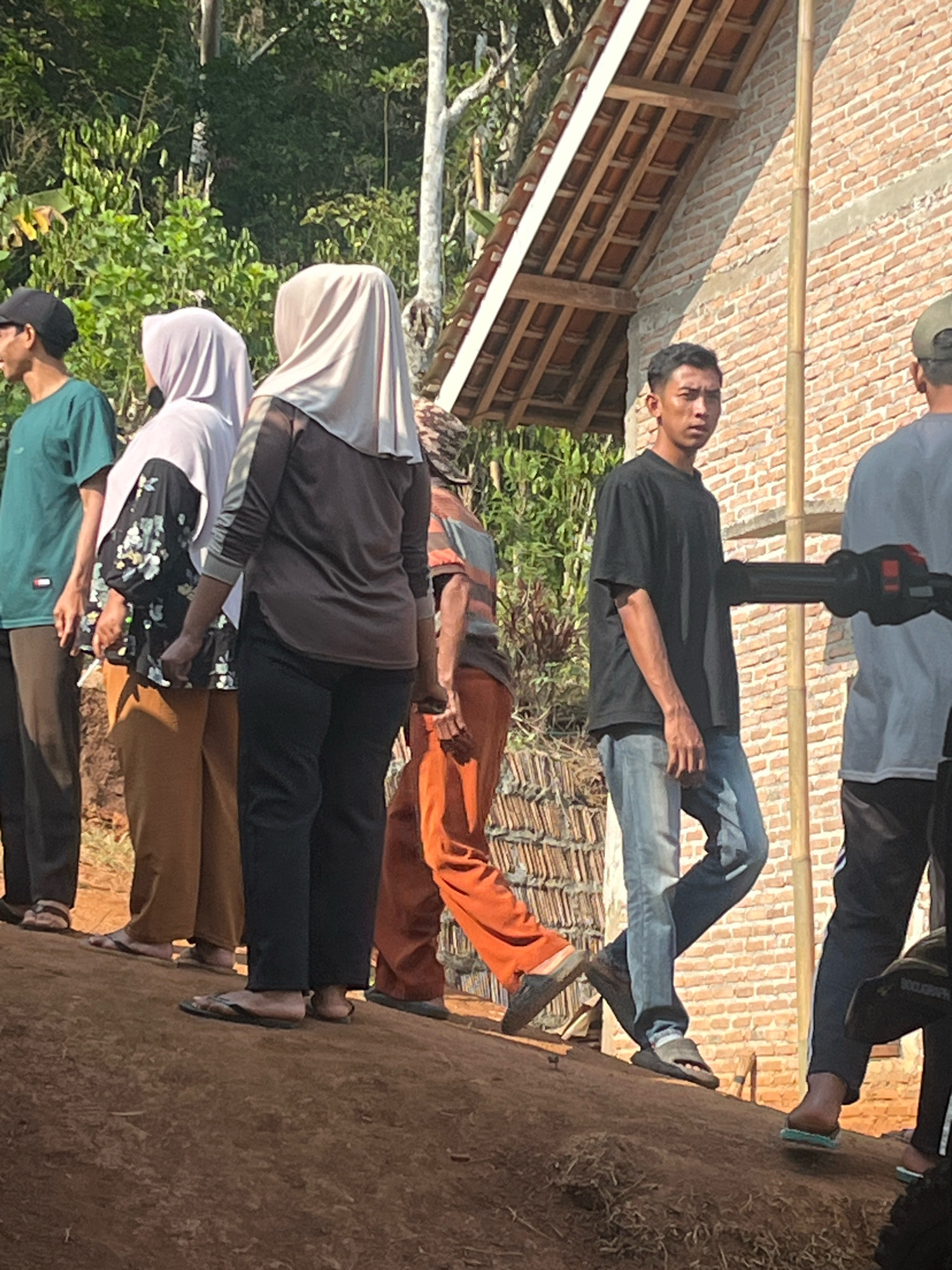 The image depicts a group of individuals, likely in Sidomulyo, Indonesia, gathered outdoors on a sloped, dirt-covered surface. The environment suggests a rural or developing area, with a brick building under construction in the background, featuring a tiled roof and bamboo scaffolding. Lush green foliage is visible behind the building.
In the foreground, several people are standing. On the far left, a person in a green t-shirt and dark pants is partially visible. To their right, two women are facing away from the camera, both wearing headscarves. The woman on the left has a light purple headscarf and a patterned blouse with black sleeves. The woman on the right wears a beige headscarf and a brown long-sleeved top with dark pants. Further to the right, a person in orange pants is visible, and then a young man wearing a black t-shirt and blue jeans stands facing the camera, looking slightly to the left. To his right, another person is partially visible, wearing dark pants with white stripes.
The overall scene is naturalistic and appears to capture a moment of interaction or waiting among the group. The lighting suggests it is daytime, possibly late morning or early afternoon, given the angle of the shadows. The ground is uneven and appears to be composed of reddish-brown soil.