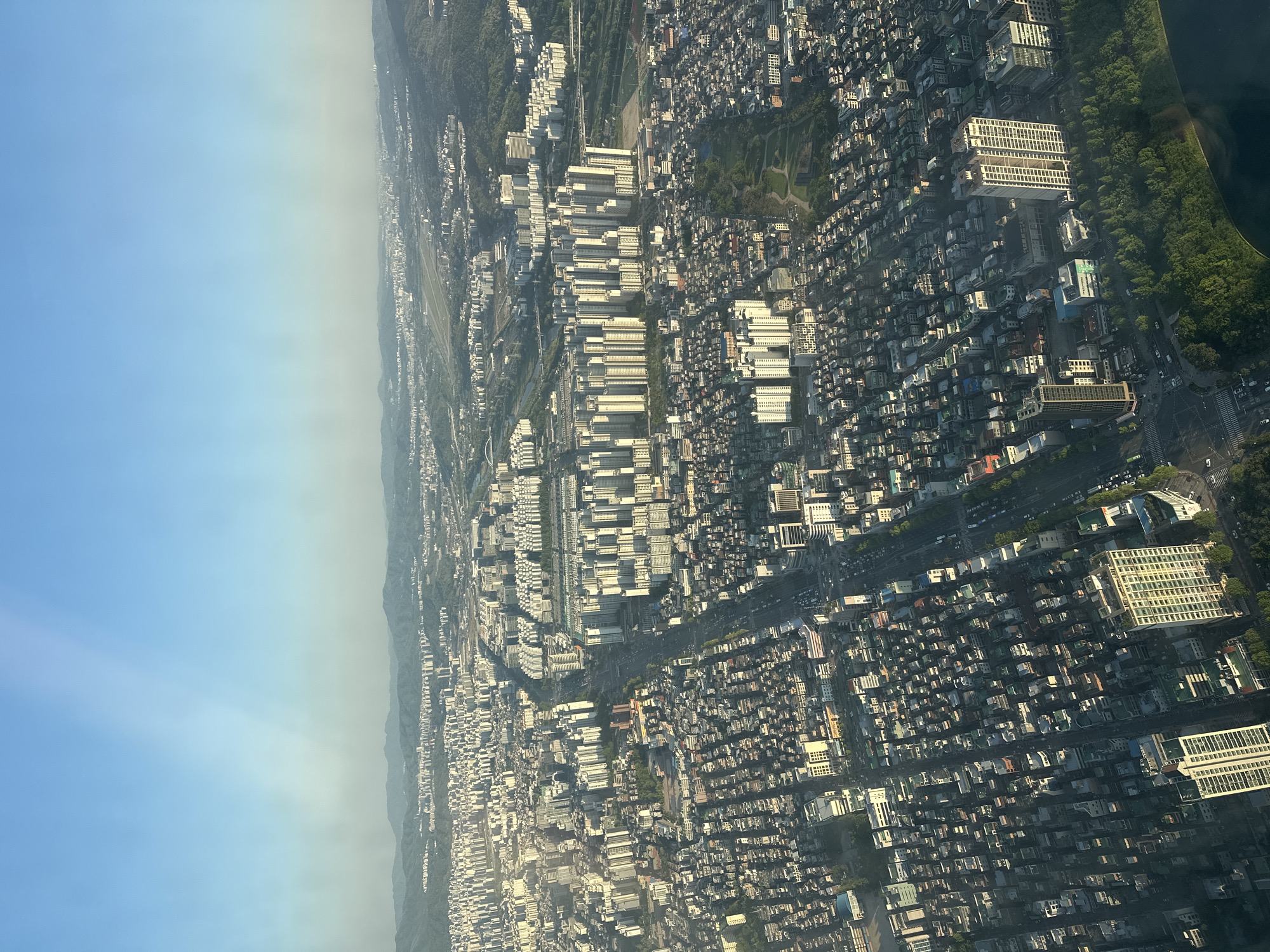 The image shows an aerial view of a densely populated urban area, likely within the city of Seoul, South Korea. Numerous buildings of varying heights are visible, from low-rise residential structures to taller apartment complexes. The buildings are closely packed together, indicative of high-density urban planning. Roads crisscross the area, with evidence of vehicular traffic. A strip of greenery, possibly a park or wooded area, is visible on one side of the urban landscape. The sky is predominantly clear with some haziness near the horizon.