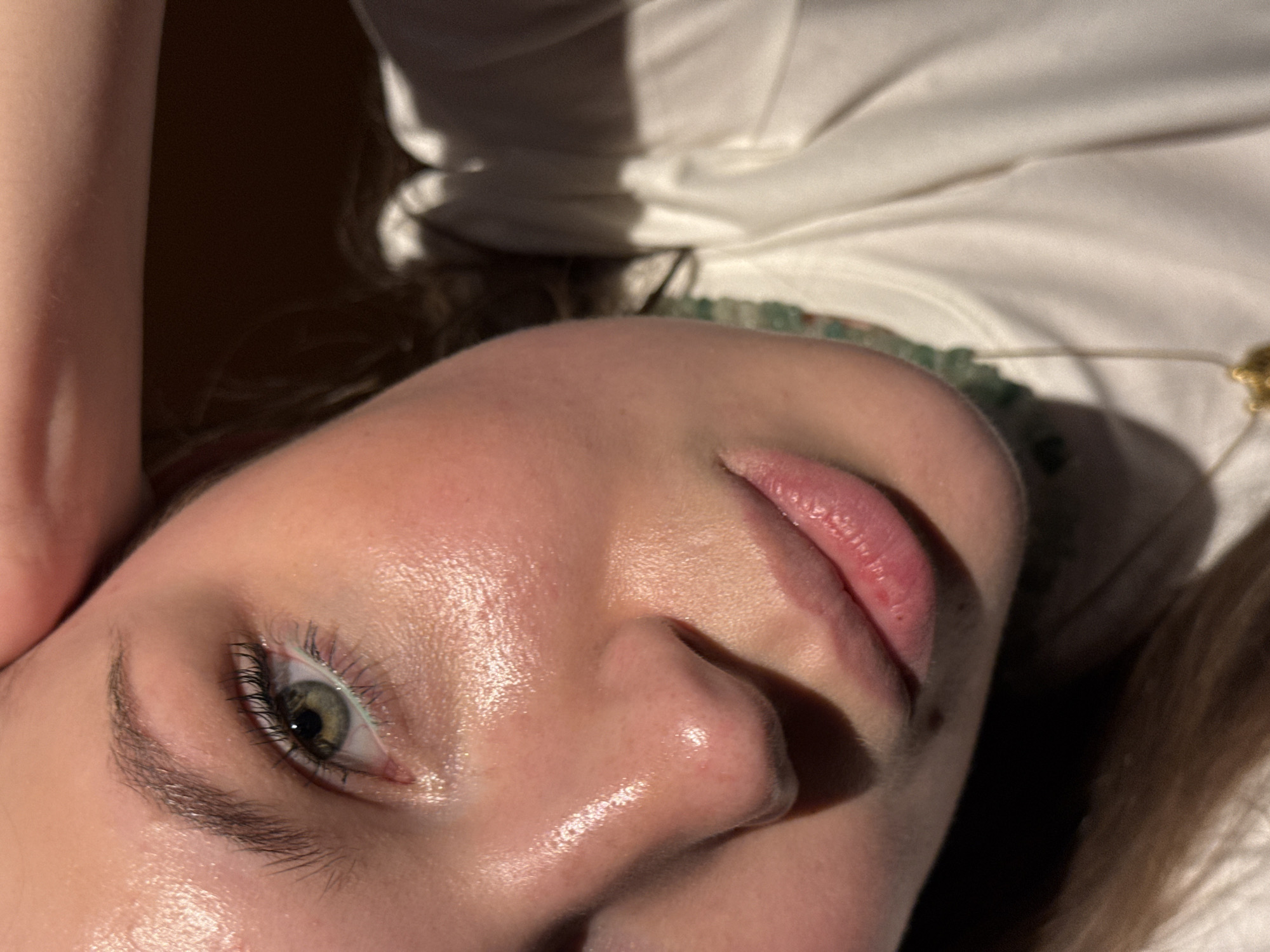 A close-up shot captures the relaxed face of a person, tilted to the side. The subject's left eye, nose, and lips are prominently featured, illuminated by soft, directional light that creates highlights on the dewy skin of the forehead, nose, and cheek. The eye is a striking green-hazel with visible eyelashes and a defined eyebrow. The lips are naturally pink and slightly parted.
The person is wearing a white garment, likely a shirt, which is visible at the top and right of the frame. Around their neck is a necklace with green, possibly beaded, elements and a small, delicate gold charm. Hair, appearing light brown or blonde, is visible at the bottom right. The background is mostly out of focus, showing dark areas alongside blurred white fabric. The overall impression is one of calm and serenity, enhanced by the natural lighting. No visible text is present. The scene is set in Belgrade, Serbia.