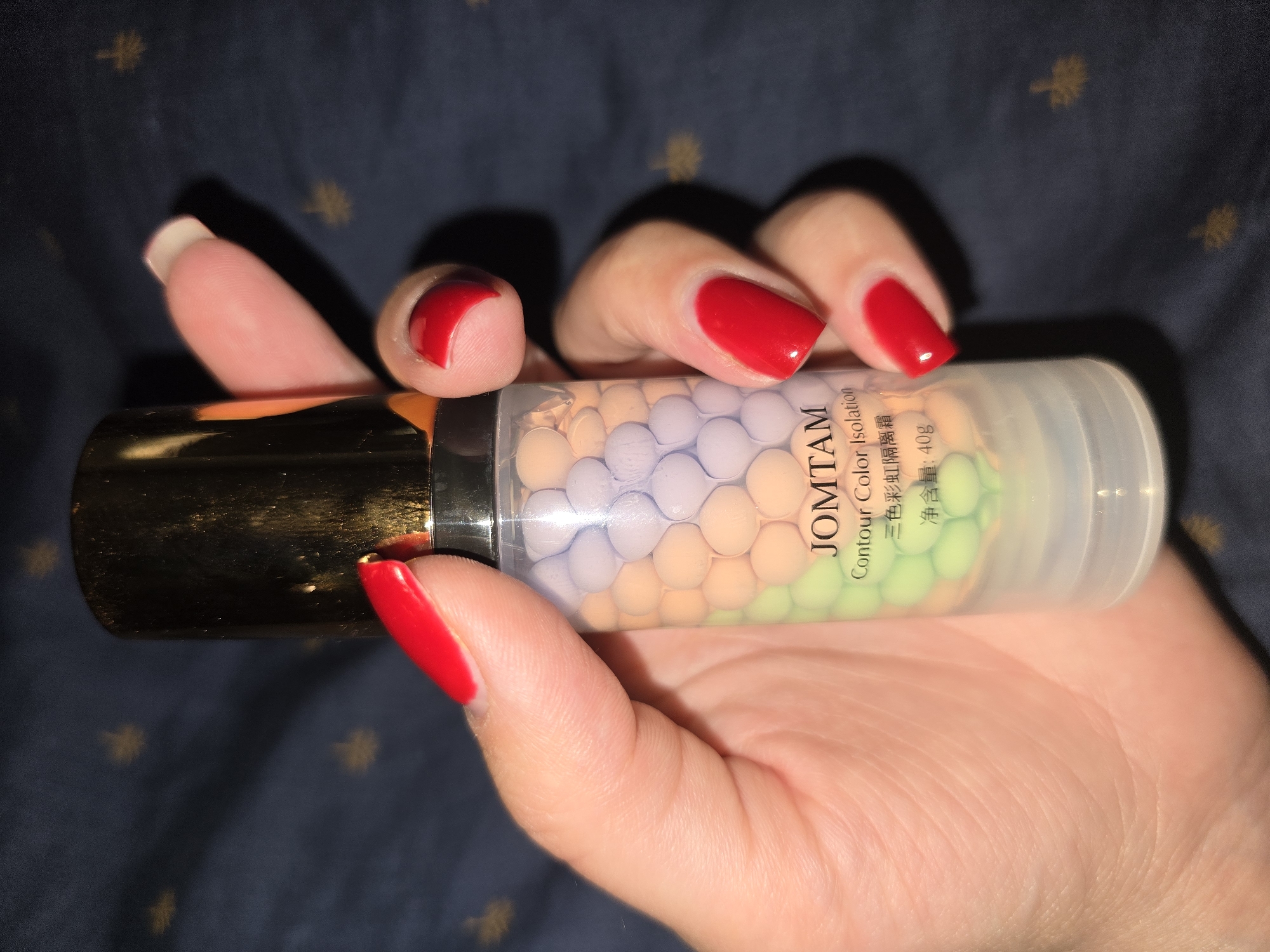 A person's hand with bright red nail polish is holding a clear plastic bottle containing small, spherical beads of various colors, including purple, peach, and green. The bottle has a gold cap and a label that reads "JOMTAM Contour Color Isolation" and Chinese characters, along with "Net Weight: 40g". The background is a dark blue fabric with a subtle pattern. The image is a close-up shot, with the hand and bottle being the primary focus. The scene is indoors, and there are no visible indicators of the location being Petropavlivska Borshchahivka, Ukraine.