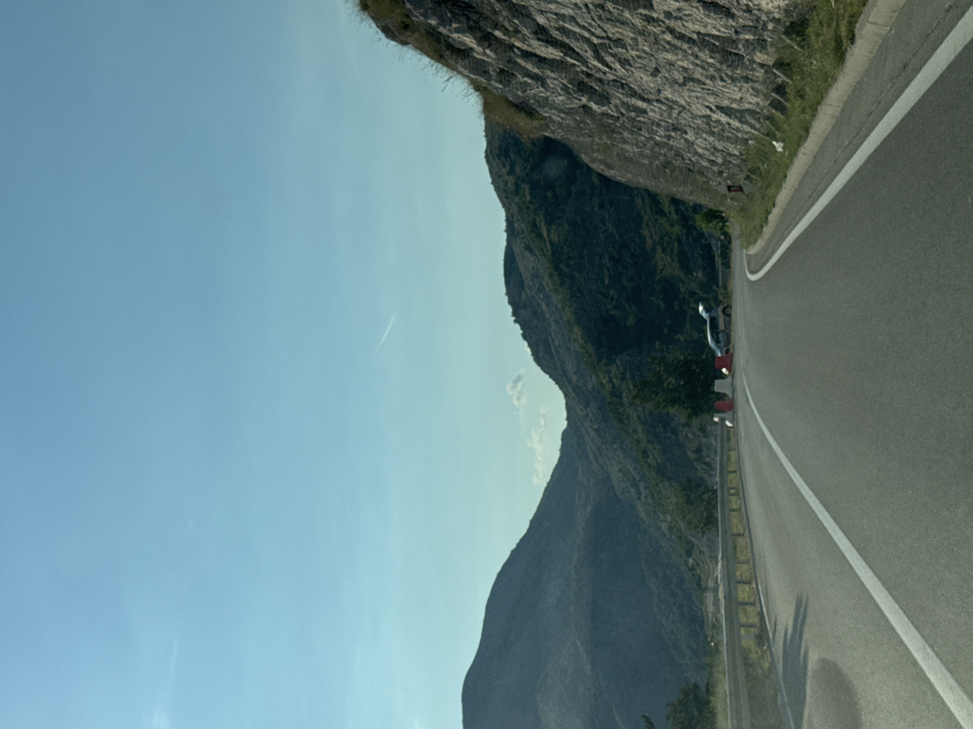 The image depicts a scenic view of a winding road through mountainous terrain, likely in or around Bukovica, Montenegro. The road, paved with asphalt, has a white line marking the center and is bordered by a metal guardrail on the left. On the right, a steep, rocky cliff face rises beside the road, covered in patches of green grass and scrub. In the background, lush green mountains stretch across the horizon, with a clear blue sky above. A few white clouds are visible, and a faint contrail streaks across the sky. Two vehicles are present on the roadside in the distance: a small silver car with its trunk open and a white van. Several orange and white traffic cones are also visible, suggesting roadwork or a temporary closure. The scene is bathed in natural daylight.