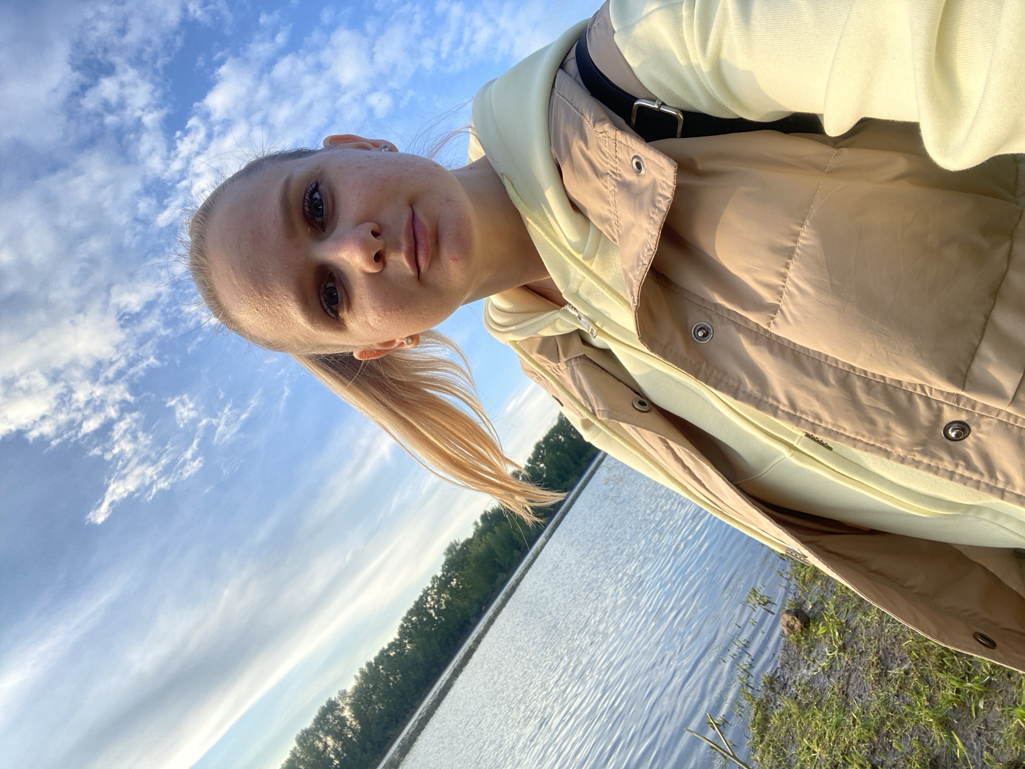 A blonde woman is taking a selfie at a lake in Chaikovsky, Russia. The sky is blue with scattered white clouds. Trees line the opposite shore of the lake. The woman is wearing a light yellow hooded sweatshirt and a tan jacket. A black strap is visible over her shoulder. The ground is grassy near the lake edge.