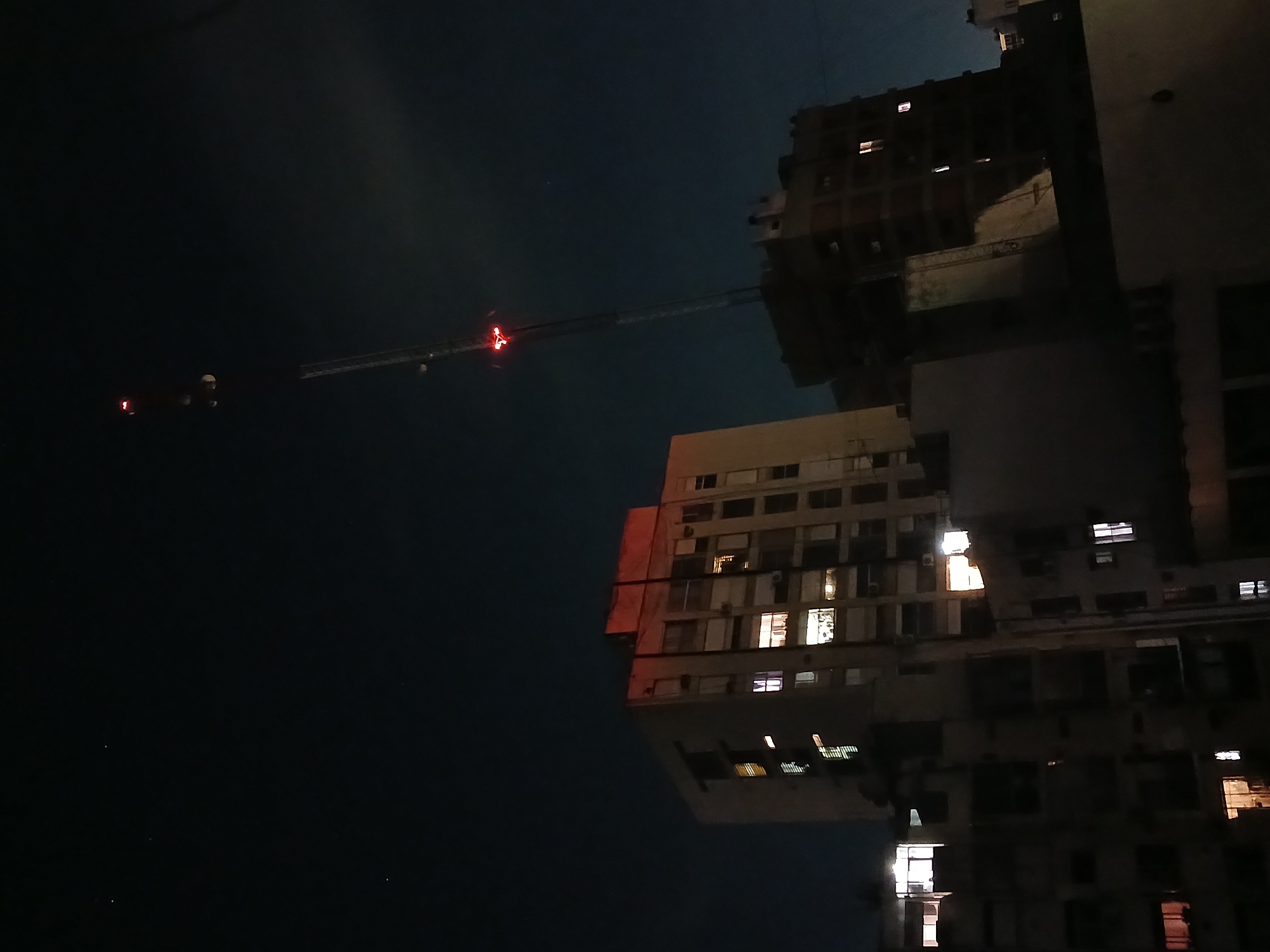 The image shows a nighttime urban scene in Buenos Aires, Argentina. Several high-rise apartment buildings are visible, with some windows illuminated. A construction crane with a red warning light extends towards one of the buildings. In the distance, there appear to be lights that could be from a vehicle or another structure. The sky is dark.