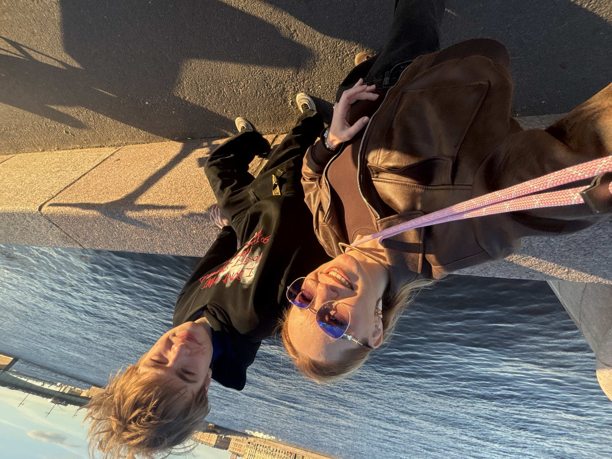 The media file shows two young adults, a man and a woman, posing for a selfie on a sunny day by a body of water, likely a river or canal, in Saint Petersburg, Russia. The man, on the left, has blond hair and is wearing a black hoodie with red lettering. The woman, on the right, has blond hair, is wearing sunglasses with reflective lenses, and a brown leather jacket over a brown top. They are both smiling, suggesting a happy and casual atmosphere. A pink leash is visible, indicating they may be with a dog, though the dog is not pictured.
The background shows a paved walkway with shadows cast by the sun, suggesting it is late afternoon or early evening, as indicated by the warm, golden light. In the distance, across the water, a cityscape with buildings is visible, consistent with a location in Saint Petersburg. The water is calm with gentle ripples. The overall impression is a relaxed, enjoyable moment shared between the two individuals. The text "I DON'T" is visible on the man's hoodie.