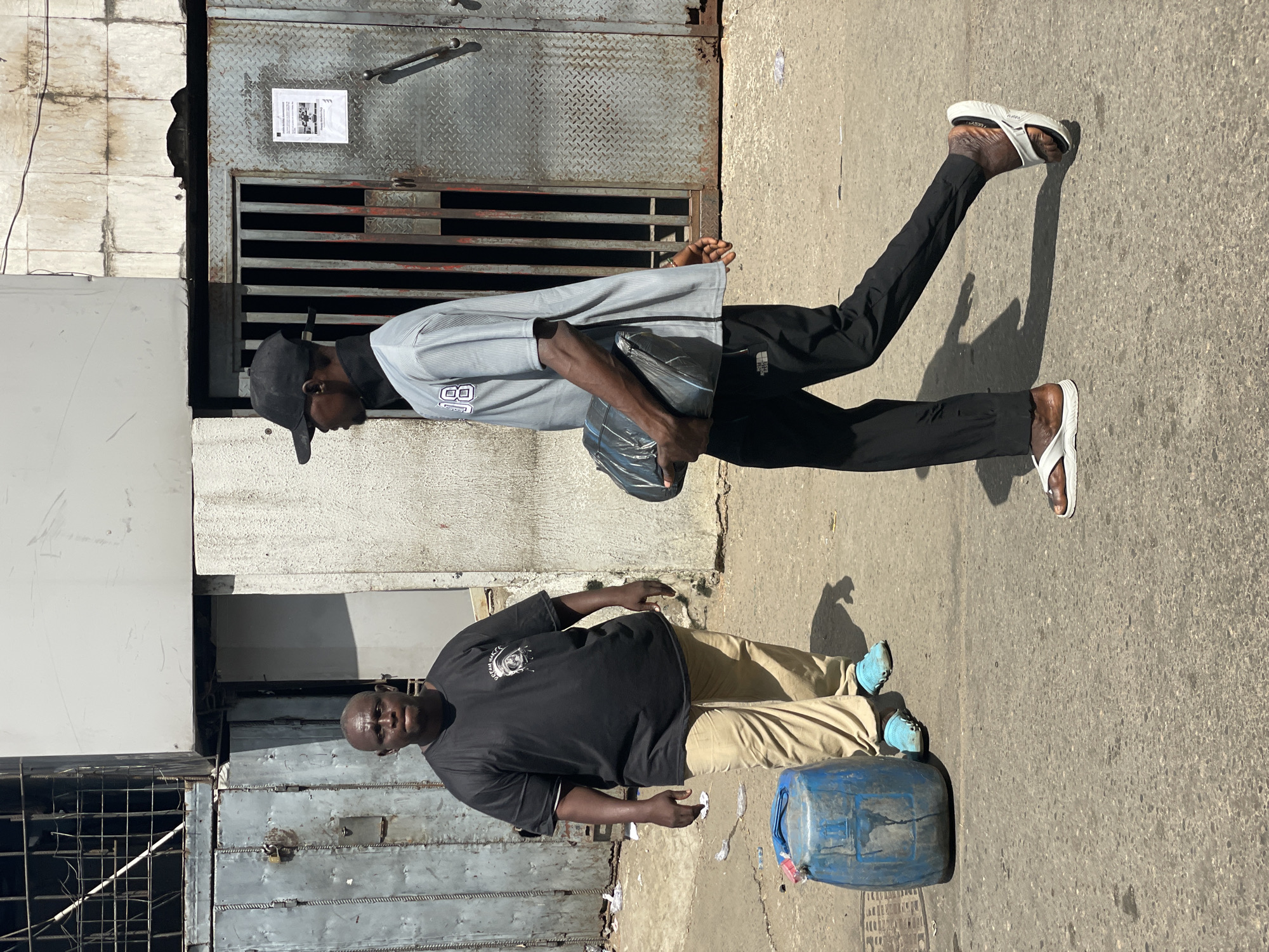 A street scene in Lagos, Nigeria depicts two men standing in front of metal structures. The first man, on the left, wears a black t-shirt and khaki pants, with blue shoes. He is standing next to a blue plastic container. The second man, on the right, wears a black cap, a gray shirt with an "8" on it, black pants, and white flip-flops. He is holding a black plastic bag. A metal door with a notice is visible behind him. The setting appears to be a bustling urban environment.