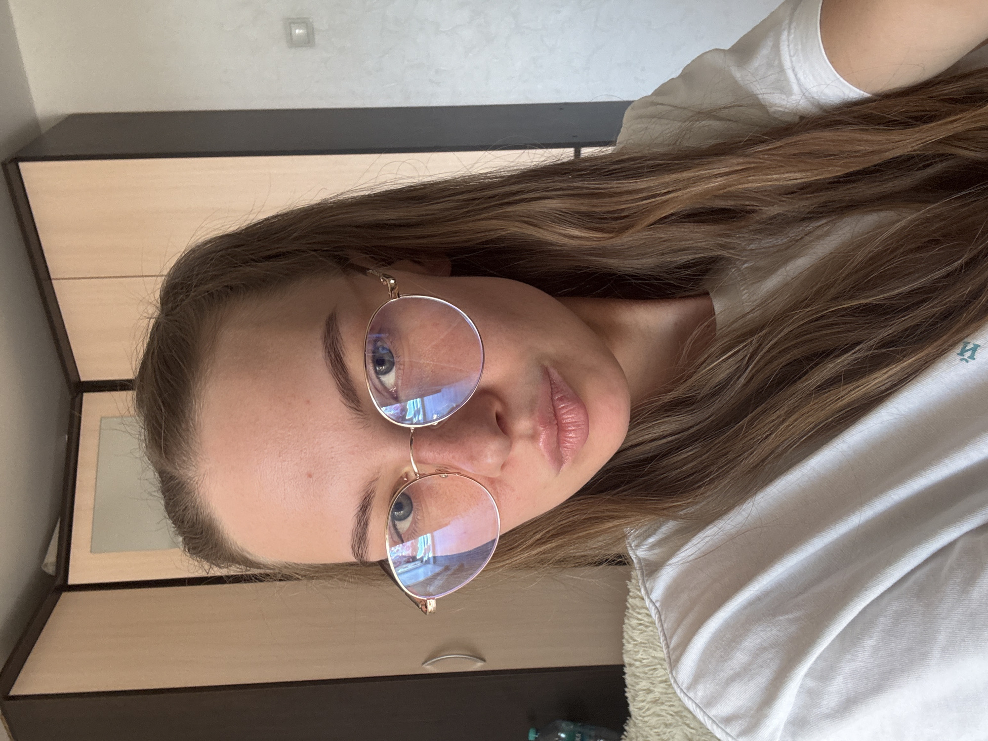 In a well-lit indoor environment, a young woman with long, light brown hair is captured in a close-up selfie. She is wearing round, gold-rimmed glasses and a white t-shirt. Her facial expression is neutral, with her lips slightly pursed. Behind her, a wooden wardrobe with light-colored doors and dark brown trim is visible. To the right of the wardrobe, a plain light-colored wall with a square light fixture is present. The overall impression is of a casual, personal moment in a domestic setting. The scene does not provide any specific indicators of its location within the city of Perm, Russia.