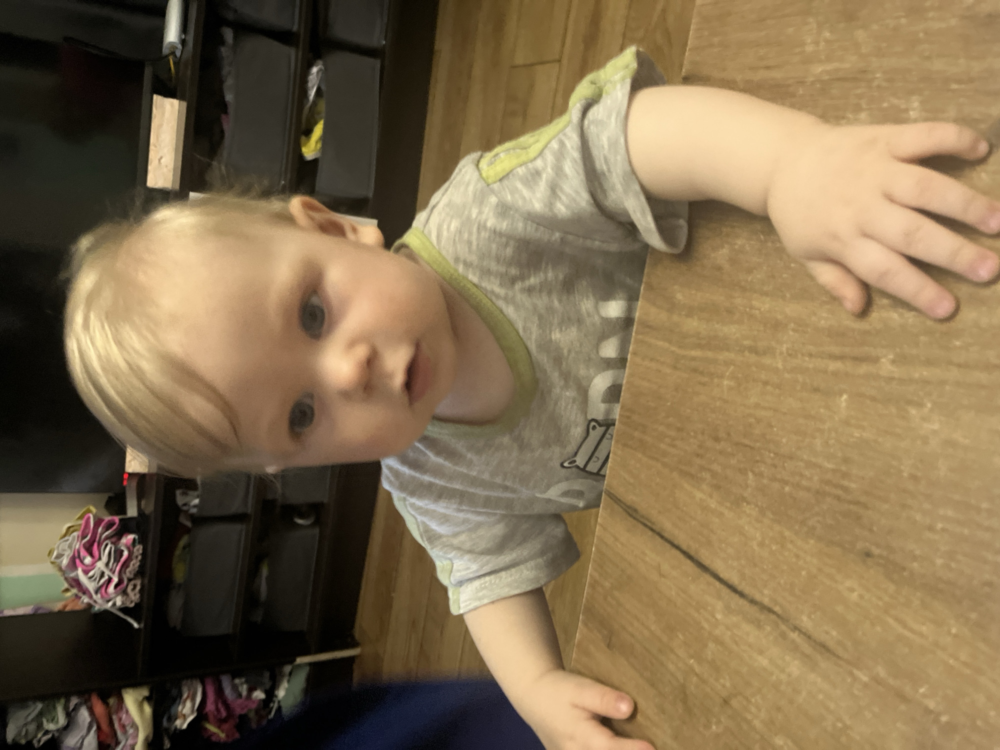 A toddler with blond hair and blue eyes is looking towards the camera. He is wearing a gray t-shirt with a green collar. His right hand is on a light brown wooden table. Behind the toddler is a black shelving unit with items stored in the shelves. The environment doesn’t provide sufficient details to confirm the city of Oryol-Izumrud, Russia.