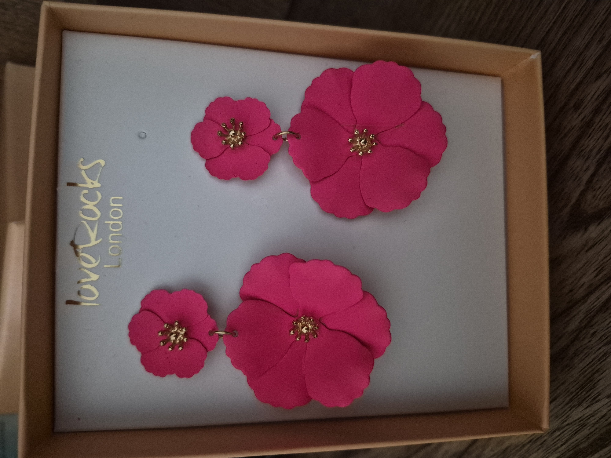 The image shows a pair of bright pink flower-shaped earrings presented in a light gray box with a gold border. The earrings are arranged in two pairs of different sizes, with a smaller flower on the left and a larger flower on the right in each pair. Both flowers have a detailed gold center. The box has gold text on the left side that reads "loveRocks London". The box is placed on a wooden surface. The lighting suggests an indoor setting, possibly a retail display or a personal collection. There are no people or specific activities depicted. The focus is solely on the jewelry and its packaging.