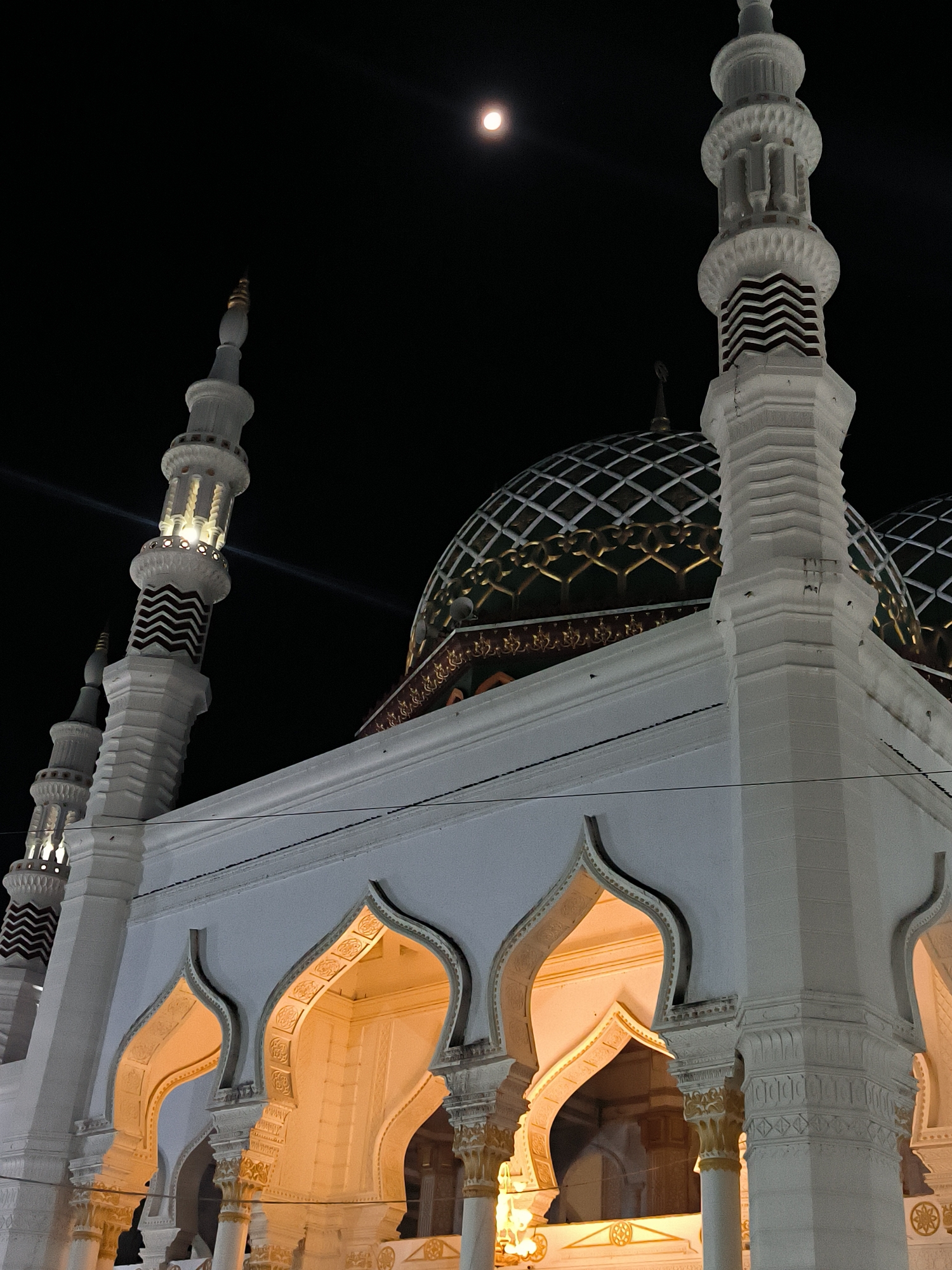 A low-angle nighttime shot shows a white mosque illuminated with warm light in Kotapantonlabu, Indonesia. Arched entrances form a colonnade, with the interior of each arch also illuminated. On top of the building are two green and gold domes. Ornate white minarets frame the dome and appear at the sides and corners of the building. At the top left of the image, the moon is visible. Dark power lines run horizontally across the image.