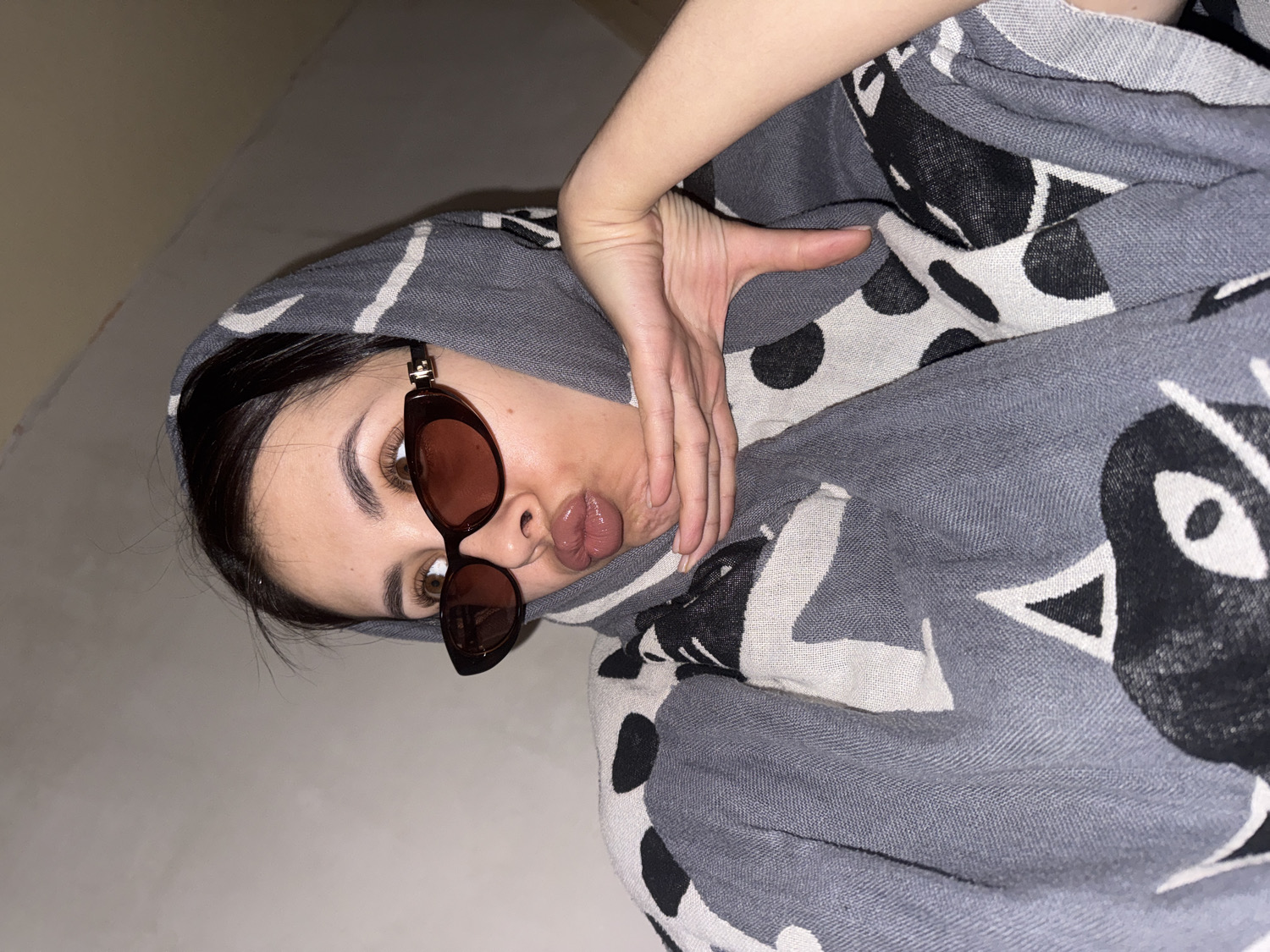 The image shows a young woman captured in a medium close-up, oriented diagonally, posing indoors in Votkinsk, Russia. She has dark hair, dark eyebrows, and brown eyes, partially visible above dark-framed sunglasses with amber-tinted lenses. Her lips are conspicuously pursed, forming a "fish face" expression. She is draped in a large, thick fabric, likely a scarf or blanket, which is patterned with a gray base color and abstract designs in black and off-white. These patterns include stylized shapes resembling eyes or animal faces, as well as polka dots. Her right hand is visible near her chin and neck, positioned as part of her pose. The background is a plain, light-colored surface, possibly a wall or floor. The lighting suggests an indoor setting, likely artificial. There is no visible text in the image. The overall impression is a casual, playful snapshot.