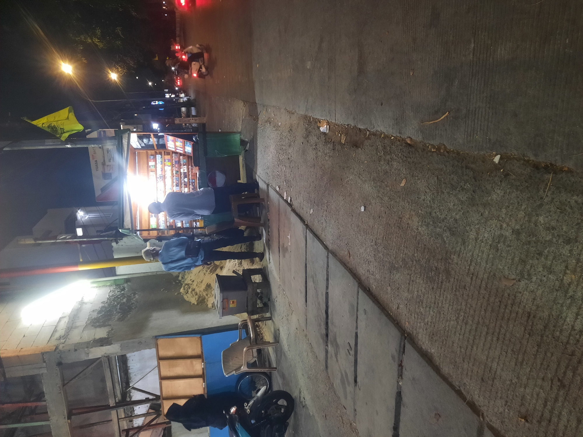 The image shows a street scene at night in North Jakarta, Indonesia, likely near a commercial area. In the foreground, a sidewalk made of concrete slabs is visible, with some debris on the ground. To the left, a person in a black hooded jacket stands near a motorcycle. Further down the sidewalk, a faded plastic chair and a dark-colored storage bin are present.
In the mid-ground, a small street stall illuminated by bright lights displays various products, possibly cigarettes or snacks, on shelves. Two men are standing near the stall. One man, wearing a light blue long-sleeved shirt and dark pants, is facing away from the camera and appears to be interacting with the stall. Another man, also wearing a light blue shirt and dark pants, is standing slightly behind the first man.
The background shows a dimly lit street with trees lining the side. Streetlights are on, and a few vehicles, including a scooter with a rider, are visible on the road, suggesting ongoing traffic. A yellow flag is attached to a pole near the street stall. The overall atmosphere is that of a typical evening in a busy urban environment.