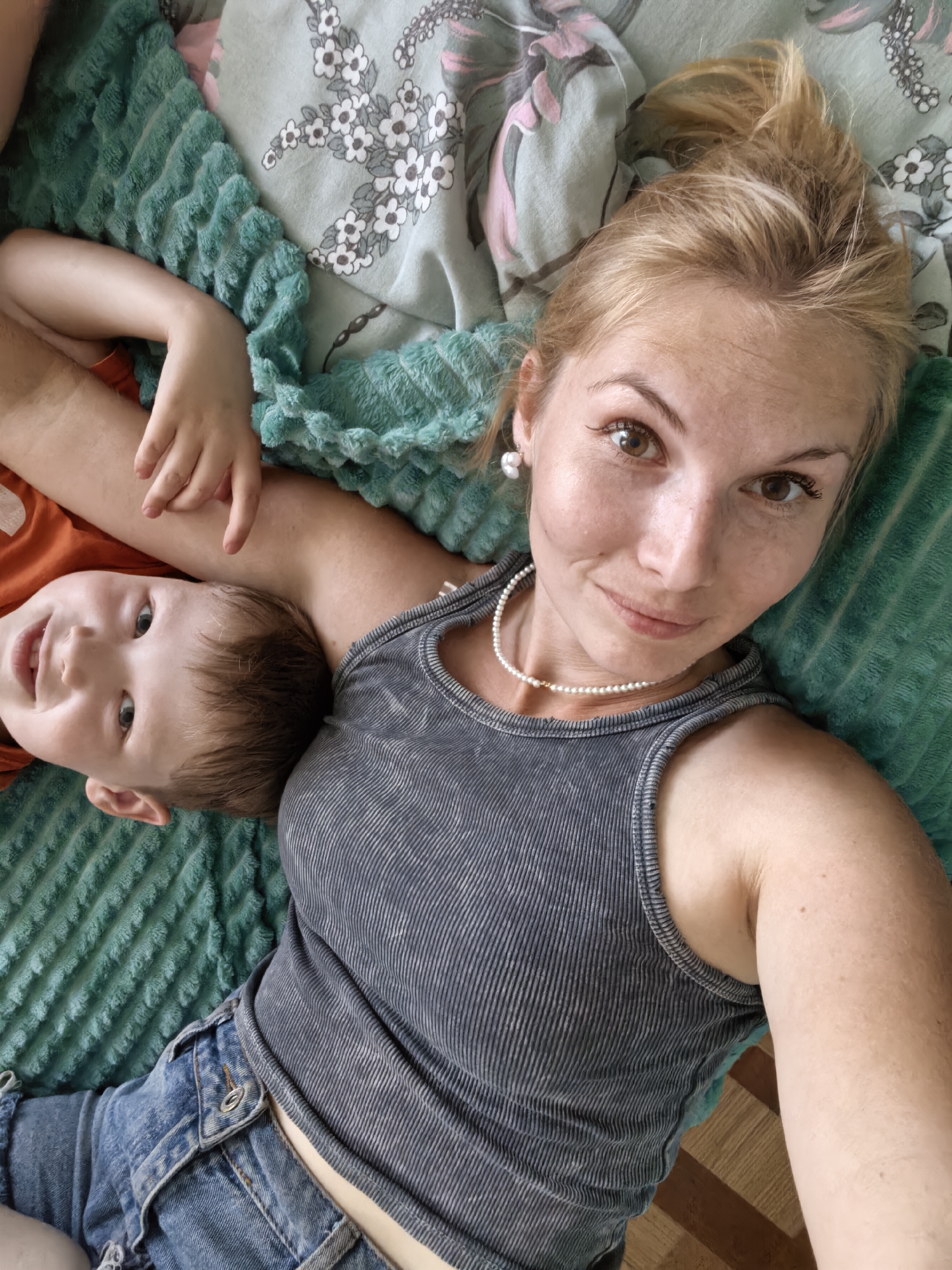 A medium shot shows a woman and a young boy lying down. The woman is on the right, looking at the camera, and the boy is on the left, also looking at the camera. They are on a teal fuzzy blanket with a floral patterned sheet underneath. The woman has blonde hair pulled up in a messy bun, brown eyes, and is wearing a grey ribbed tank top and a pearl necklace. The boy has brown hair and is wearing an orange t-shirt. His left arm is around the woman's neck and his right hand is on her arm. The woman's left arm is around the boy. The image is taken from a high angle.