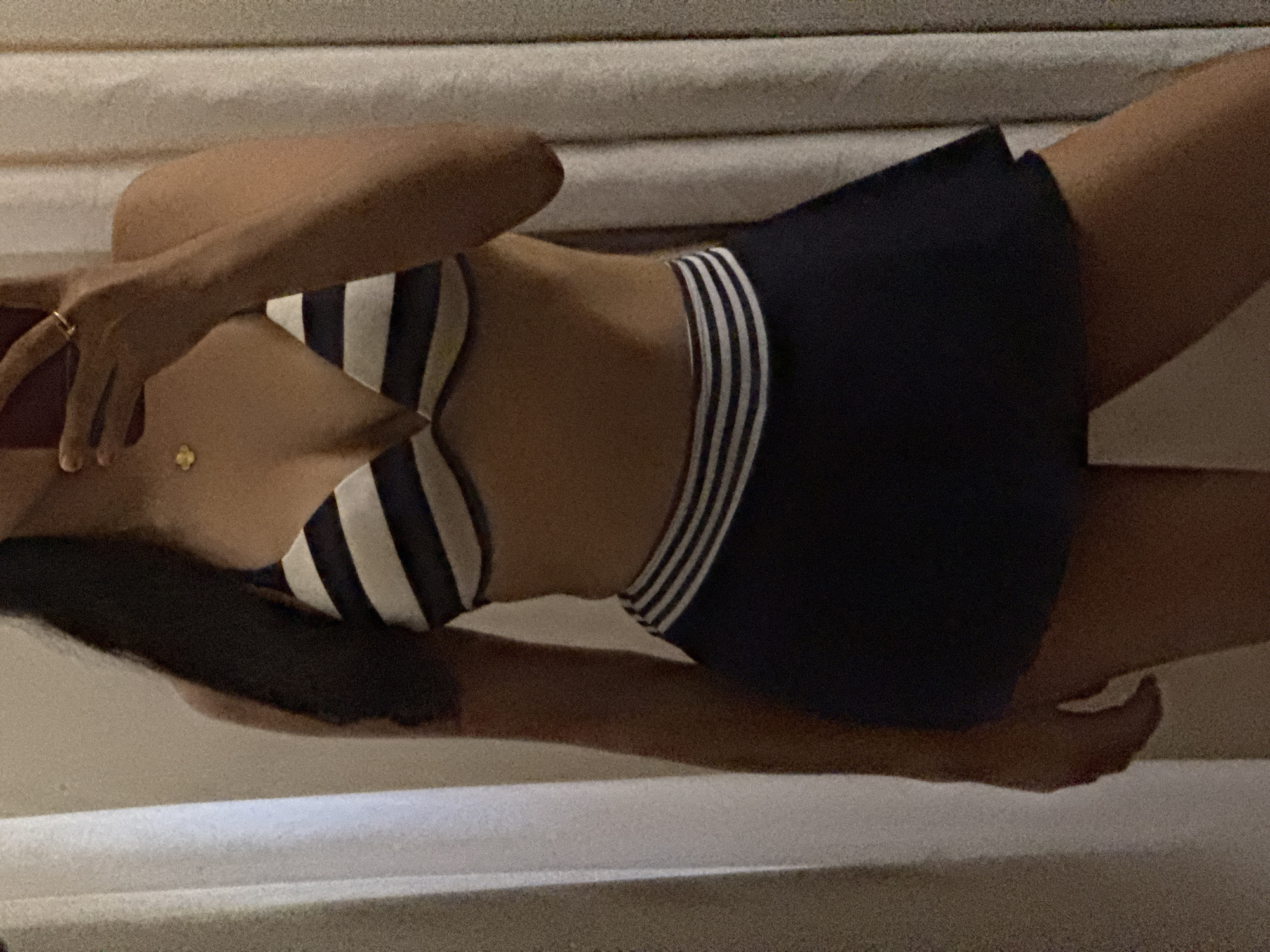 A female individual stands indoors, captured in a mirror selfie. Her dark hair is long and falls over her left shoulder. She wears a two-piece swimsuit set consisting of a striped bikini top with horizontal black and white bands, and a dark navy or black swim skirt with a waistband featuring multiple thin white horizontal stripes. A small, gold-colored four-leaf clover necklace is visible on her chest, and a gold-colored ring is on the index finger of her right hand, which holds a red-cased smartphone in front of her face. The background includes light-colored walls and beige or off-white curtains on both the left and right sides of the frame.