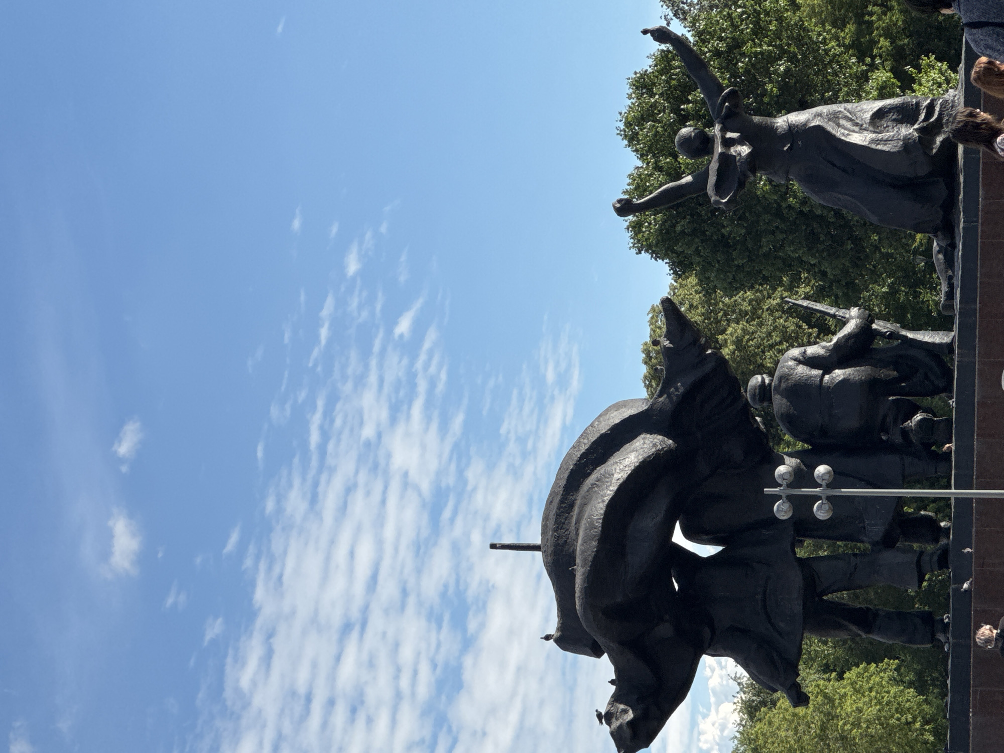 A black bronze sculpture group stands in a park-like setting with a bright blue sky and scattered white clouds. The sculpture depicts several figures, including one woman with arms raised in a gesture of triumph or supplication, kneeling on a flowing gown. To the left, other figures are partially obscured, carrying what appears to be a large banner or cloth. Birds are perched on various parts of the sculpture. Trees form a dense green backdrop behind the artwork. In the foreground, a few people are visible, and a lamppost with four globes stands near the center. The sculpture is located at Victory Square in Moscow, Russia, a prominent landmark commemorating the Soviet victory in World War II.