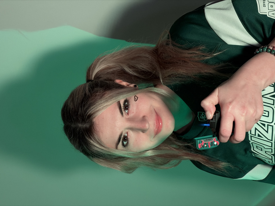 The scene is situated in Budva, Montenegro. The image depicts a young adult female, approximately mid-20s, with light blonde and brown hair styled in two pigtails. She has light skin, brown eyes, and a small black heart drawn below her left eye. She wears small black stud earrings. Her lips are glossy, and she exhibits a faint, closed-mouth smile while looking directly at the viewer. She is dressed in a dark green long-sleeved jersey with white accents on the sleeves. The word "ARIZONA" is prominently visible in white text on the front of her shirt, and a stylized "AA" logo is present near the collar. Partially visible on the lower right, the text "OFFICE OF ARMY" can be discerned on her shirt. On her right wrist, she wears a multi-colored beaded bracelet. Her right hand holds a black lighter, which is ignited, producing a distinct blue flame. The lighter's lid features a green and red cartoon-like dinosaur design. The background consists of a plain wall, uniformly illuminated with a strong green light, creating visible shadows from the subject's head and shoulder on the right side of the frame.