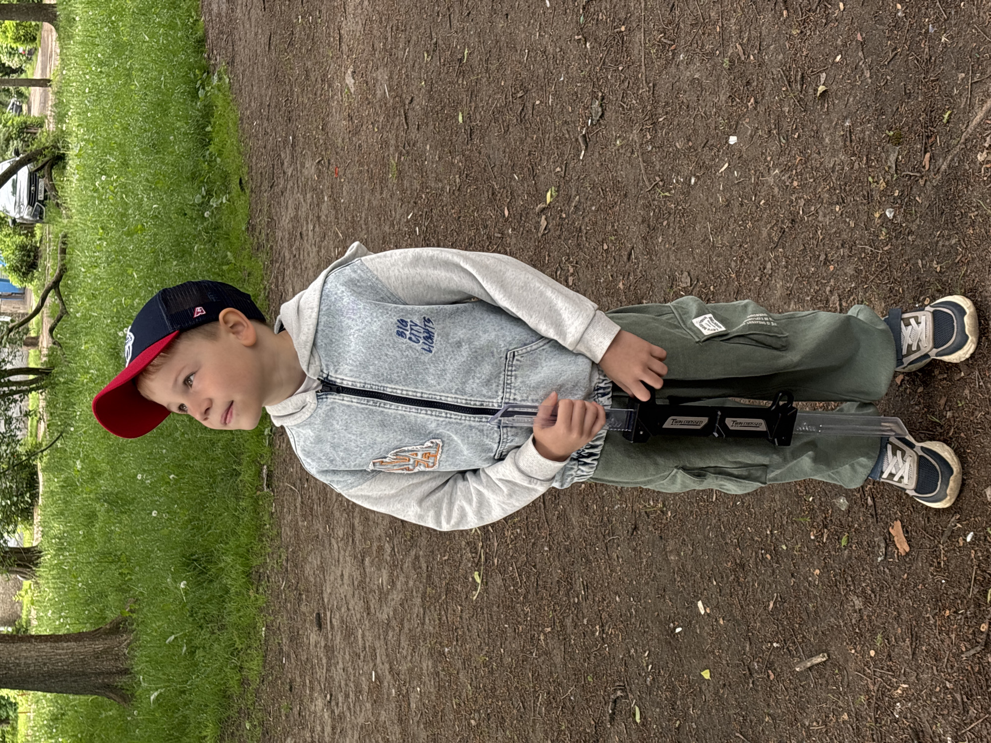 A young boy is captured in an outdoor setting, standing on a dirt path. The image is oriented sideways, but he is positioned upright, looking towards his left. He wears a navy blue and red baseball cap, a light gray long-sleeved hooded shirt under a light blue denim vest-style jacket, dark green cargo pants, and blue and white sneakers. His hands hold a clear and black toy, possibly a blaster or water gun, horizontally in front of him.
The path beneath him is earthy brown, with scattered debris and small green shoots. To his left, a vibrant green grassy area extends, interspersed with small white flowers. Trees with visible trunks are present in the grassy section, and further in the background, a light-colored car is partially visible amongst the foliage, suggesting proximity to a road or parking area. The lighting appears soft, indicating an overcast day.
Notable text includes "BIG CITY LIGHTS" embroidered on the front of his denim jacket. On the toy he is holding, "Twin Crossed" is visible in white lettering on a black segment in two places. A small white label on his green pants shows partially legible text, possibly starting with "LET'S EXP..."