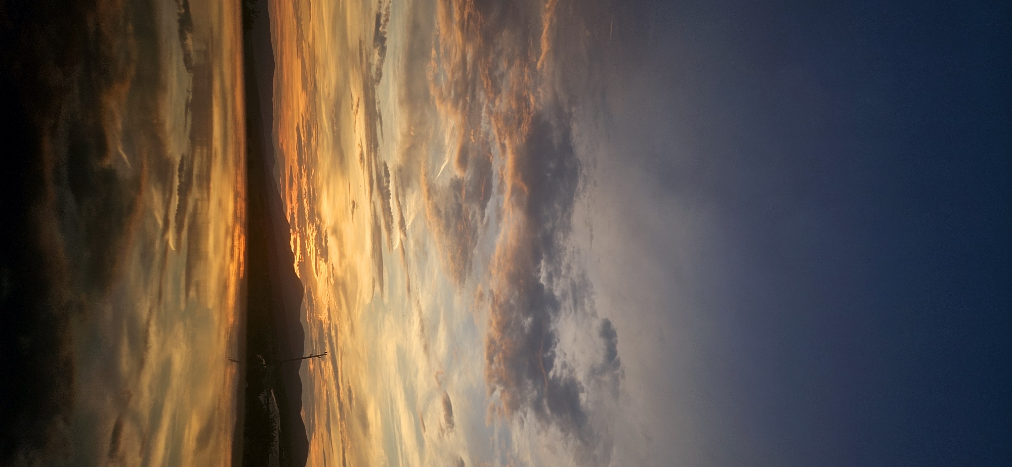 This image captures a serene sunset over a body of water, likely a lake or river, near the town of Momin Prohod, Bulgaria. The sky is a gradient of deep blue at the top, transitioning to shades of orange, pink, and yellow as it nears the horizon, painted with wispy clouds. The warm hues of the sunset are beautifully reflected in the calm water, creating a mirror image of the sky and the silhouetted mountain range in the distance. A thin, dark line of mountains marks the horizon, with subtle indications of vegetation and possibly some man-made structures. A single, thin pole or antenna is visible on the right side, standing tall against the twilight. The overall atmosphere is tranquil and picturesque, characteristic of a peaceful evening in the Bulgarian countryside. There are no people or animals visible in the scene.