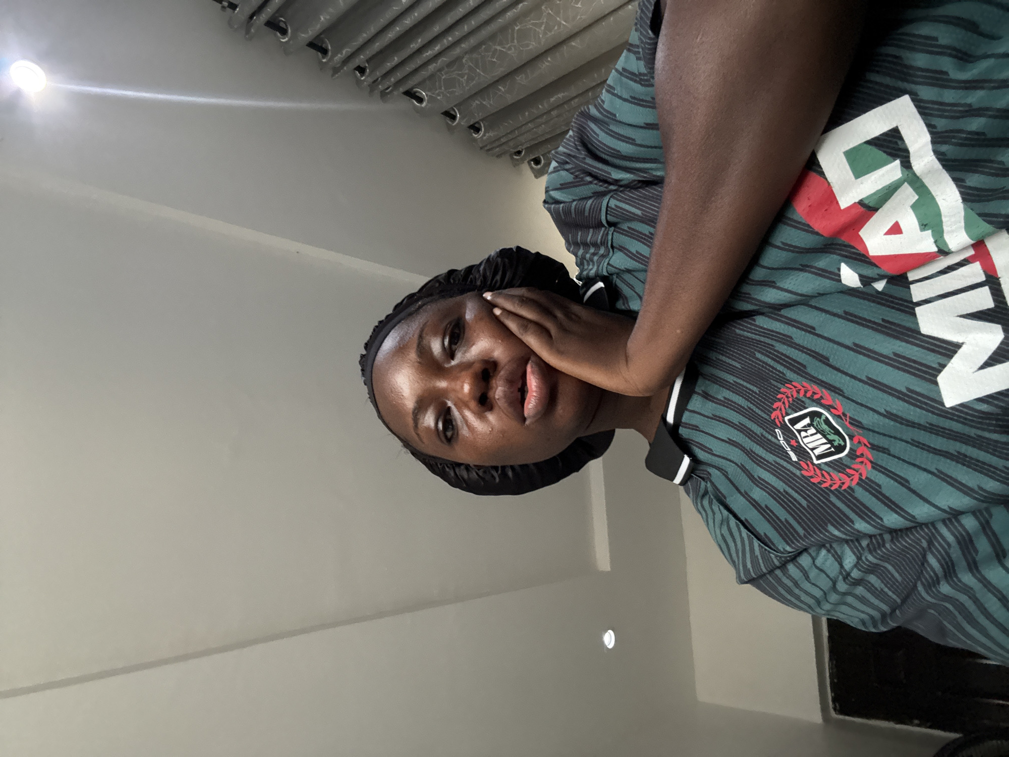 A dark-skinned woman with a black head covering is posing for a selfie indoors. She is wearing a green and black striped shirt with "Milan" printed on it and the logo of the Nigerian Professional Football League (NPFL). Her hand is on her cheek. The ceiling and curtains are visible in the background. The specific location in Iddo Tudunwada, Nigeria, cannot be determined from the image.