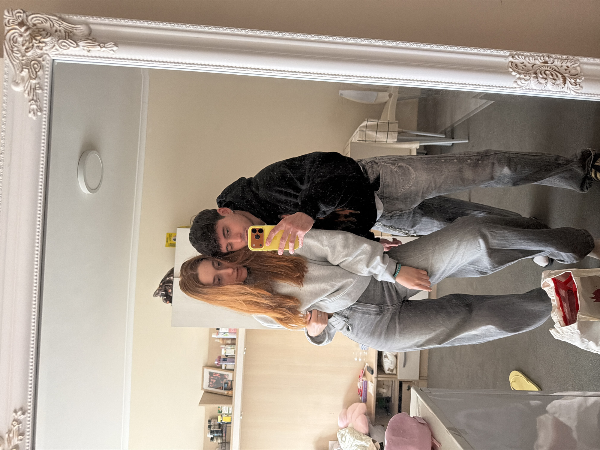 The image captures a mirror selfie of two young individuals, a male and a female, taken in an indoor setting in Sofia, Bulgaria. The photograph is presented rotated 90 degrees counter-clockwise, showing the subjects standing side-by-side in the reflection of a large, ornate white mirror with decorative carvings on its frame.
The male, positioned slightly behind the female, has dark hair and is dressed in a black hoodie and grey jeans. He is holding a yellow-cased smartphone with his right hand to capture the reflection, his arm around the female. The female, with long reddish-brown hair, wears a light grey sweatshirt and grey jeans. A turquoise bracelet is visible on her left wrist as she leans into the male. Both subjects look towards the phone's reflection with neutral to subtly smiling expressions, conveying a relaxed and affectionate mood.
The background reflected in the mirror reveals a room with light-colored walls and a grey carpet or rug. Visible objects include shelves stocked with various bottles and small items, framed pictures, a white chair or stool, and a round white light fixture mounted on a wall. On the floor, a white bag with red accents, yellow flip-flops, and pink fluffy items are discernible, suggesting a casual home environment, possibly a bedroom or living area. There is no clearly legible text within the image.