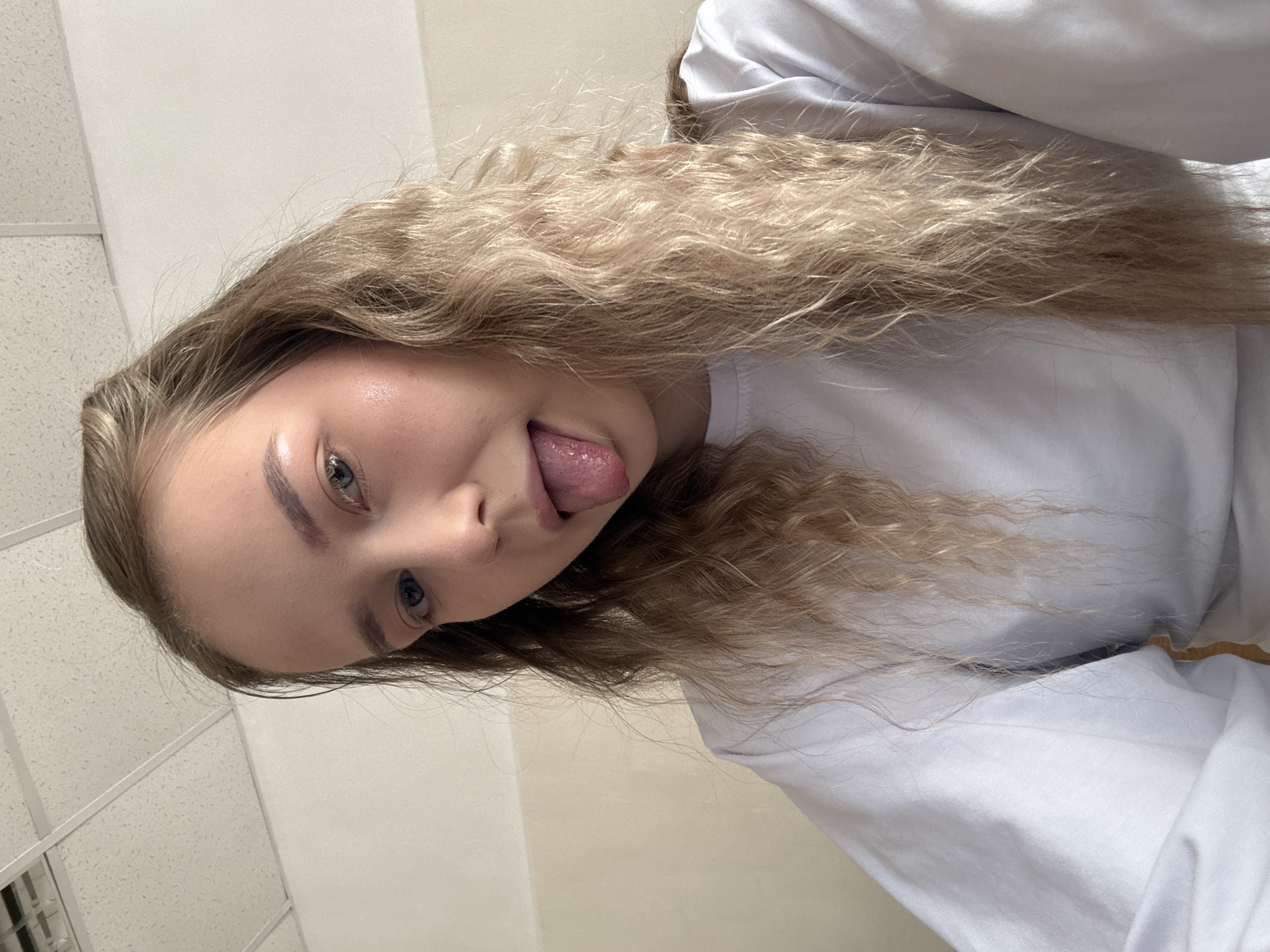 The image shows a young person in a close-up selfie, oriented on its side. The subject has long, wavy, blonde hair that cascades around their face. Their face is partially visible, featuring striking blue-grey eyes looking towards the camera, and a playful expression with their tongue slightly extended. The skin on their face appears smooth and fair, with a subtle highlight on the forehead and cheekbone. They are wearing a simple white long-sleeved top.
The background indicates an indoor setting. On one side, a plain, light-colored wall, possibly beige or off-white, is visible. On the opposite side, a white ceiling with a grid pattern, common in institutional or commercial buildings, is present, along with a ceiling panel or light fixture. The lighting is bright and artificial. The overall impression is one of youthfulness and a lighthearted moment. There is no discernible text in the image. The location context for this media file is Chaikovsky, Russia.