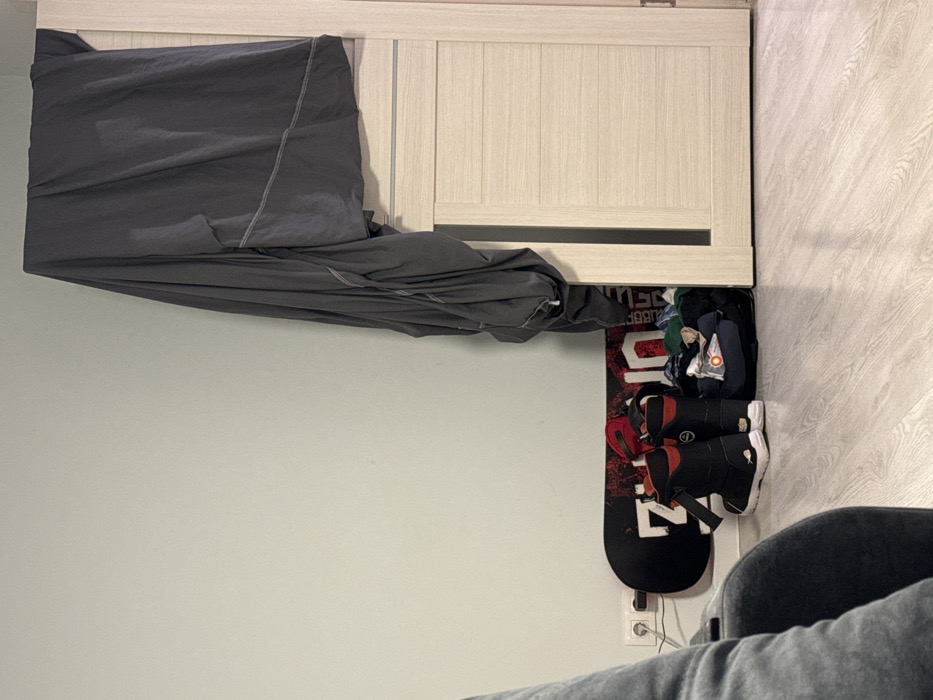 A cluttered scene displays a snowboard with attached boots standing against a light gray wall. Partially visible, a closet-like structure with beige doors and a black horizontal accent line sits above the snowboard. A dark gray cloth drapes over the top of the closet. Clutter, possibly including clothes, is also behind the snowboard. A gray chair and legs clad in gray pants occupy the bottom right corner of the image.