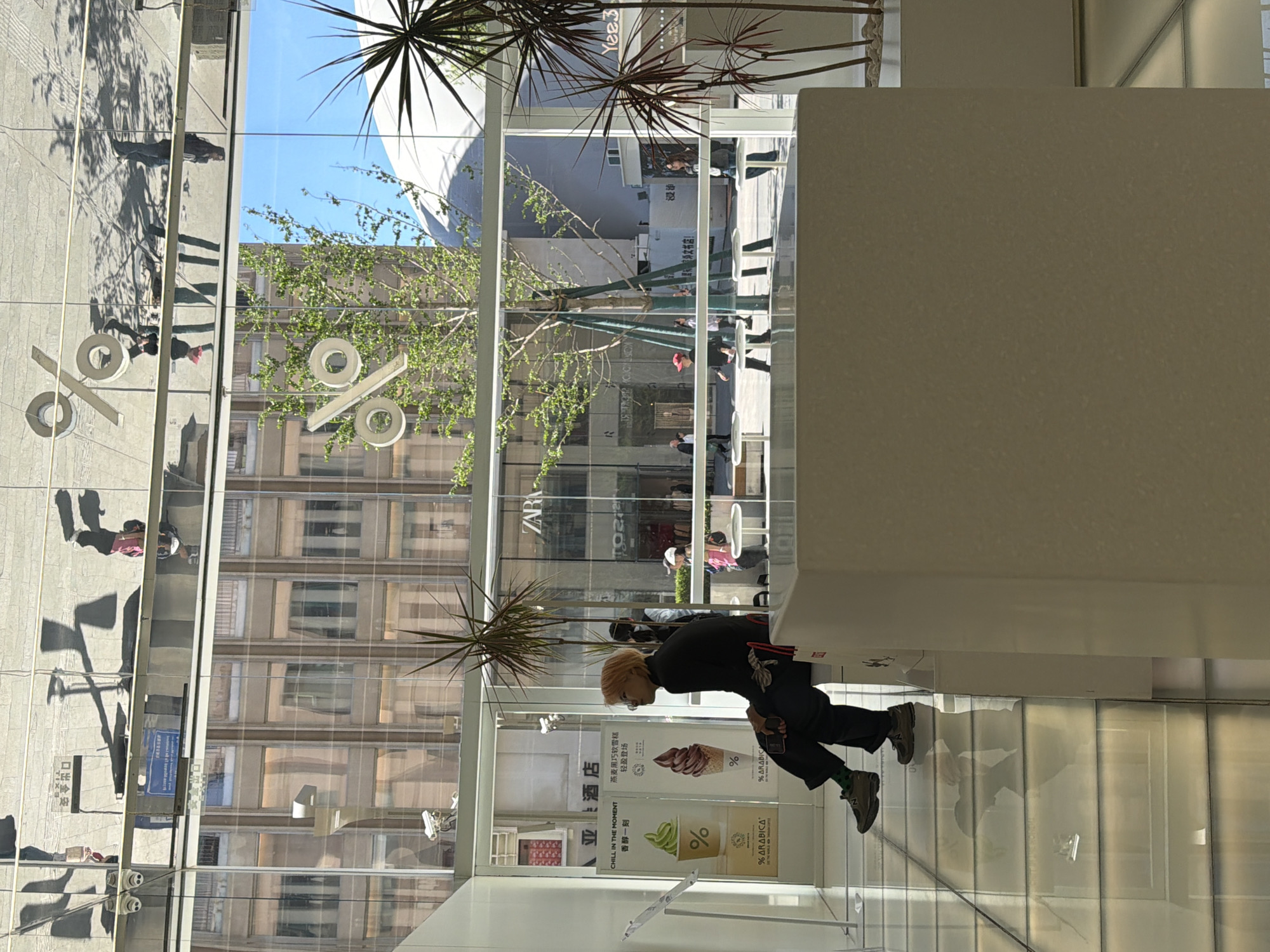 Here is a concise description of the image:
The image is taken indoors, looking outwards through a large glass window. On the left of the window, a large symbol resembling a percentage sign can be seen. On the right side, there is a person bending over, and they appear to be looking at something. Signage is visible for % ARABICA coffee shop and 亚酒店. Buildings and trees are visible through the window, as well as street activity, which suggests an urban environment. The presence of Chinese characters on the signage suggests the location is within China, likely in the Dongcheng District.