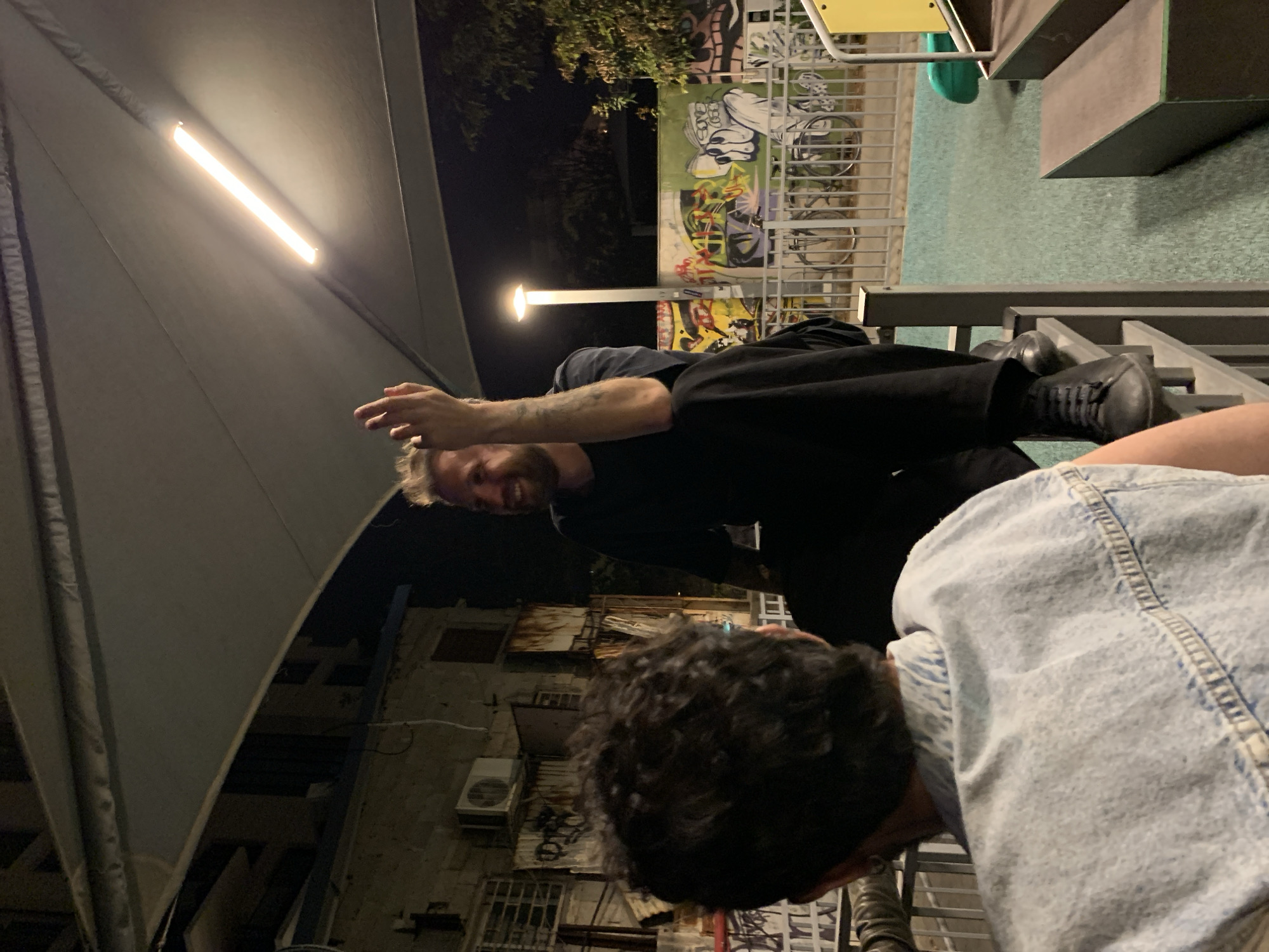 Scene: An outdoor nighttime gathering, possibly in Tel Aviv, Israel.
People: Two individuals are visible. One, with fair skin and short, blond hair, is wearing a dark shirt and black pants and shoes. His arm features visible tattoos. He seems to be gesturing toward the camera. The other person has dark hair, is wearing a light-colored denim jacket, and is facing downward.
Setting: The background features a partially visible wall adorned with graffiti, a building exterior with windows and air conditioning units, and a playground area with climbing structures and a green surface. A bright, elongated light source illuminates the foreground.
Objects: Clothing, footwear, playground equipment, graffiti, building facade, windows, AC units, light fixture.
Actions: The two individuals appear to be interacting, while one gestures towards the camera.
Environment: The lighting suggests it is nighttime. The urban environment indicates a city setting.
