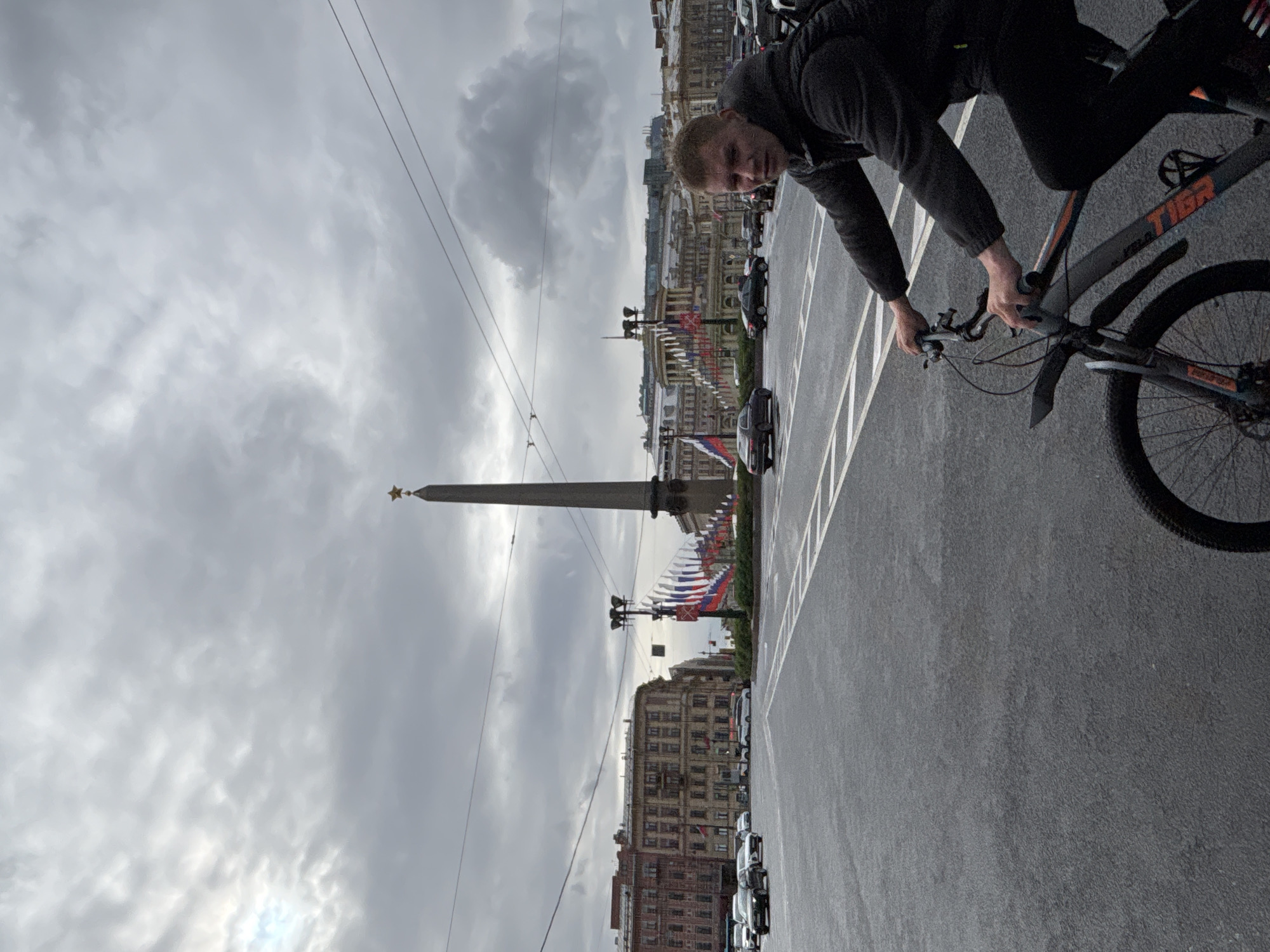 A man rides a bike on a road in Saint Petersburg, Russia, next to the Winter Palace. The road has white lane markings. The Alexander Column in the center of Palace Square is visible in the background, with Russian flags hanging from the buildings. The sky is cloudy. Several cars are parked along the side of the road.