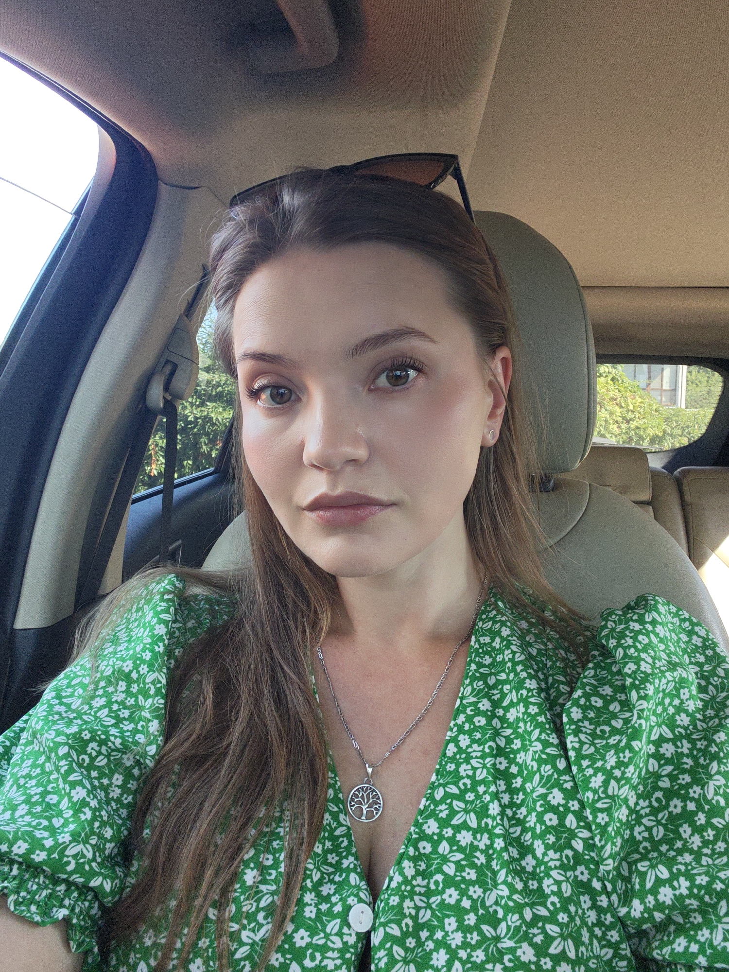 A young woman with light brown hair is sitting in the driver's seat of a car. She is wearing a green dress with a white floral pattern and a silver necklace with a tree pendant. The car's interior is visible, with the beige leather headrest of the passenger seat and the car's ceiling. Outside the car windows, green foliage and a building can be seen. The woman is looking towards the camera with a neutral expression. The image appears to have been taken in Vinnytsia, Ukraine, though no specific landmarks are identifiable.