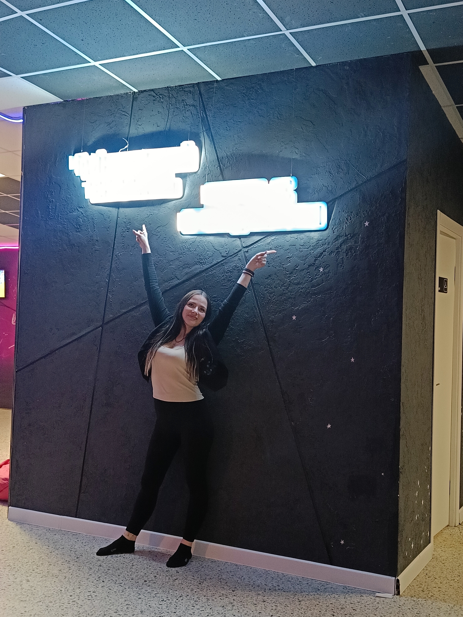 A woman is standing in front of a textured black wall with illuminated neon signs, possibly in Chaikovsky, Russia. The woman has dark hair and is wearing a white top, black leggings, and a dark jacket. She is pointing with both hands towards the neon signs. The wall is decorated with a pattern of lines and small stars. The flooring is speckled. A white door is visible to the right. The ceiling has a grid pattern.